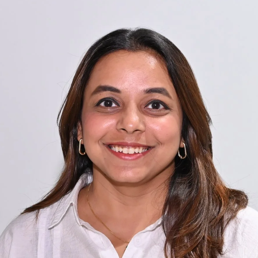 Close-up of a smiling woman with long brown hair, wearing a white collared shirt, gold hoop earrings, and a delicate gold necklace, against a plain white background.