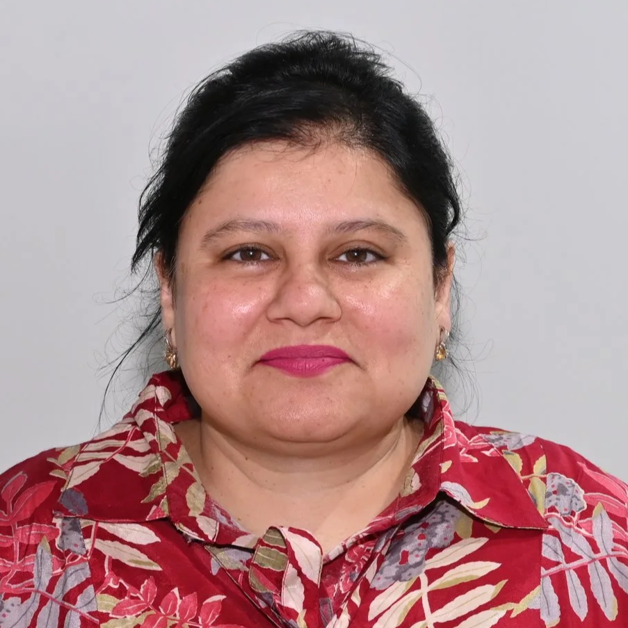 A woman with dark hair wearing a red floral patterned blouse and pink lipstick, looking at the camera against a plain gray background.