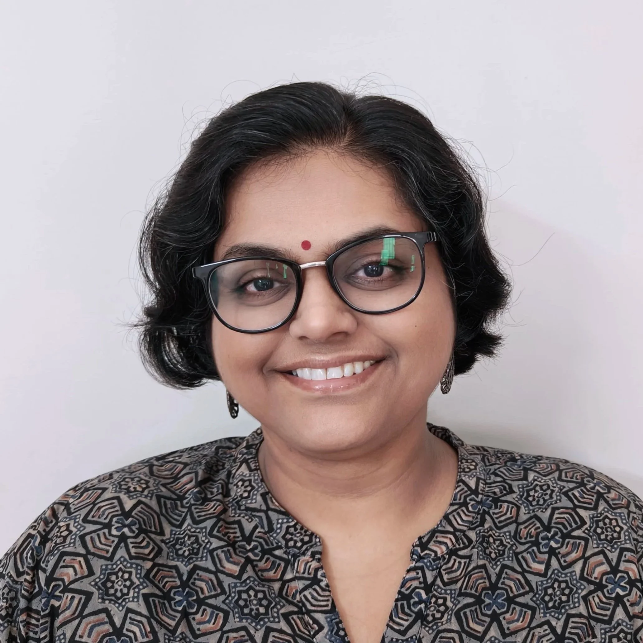 A woman with short black hair, glasses, and traditional Indian earrings wearing a patterned top, smiling against a plain white background.