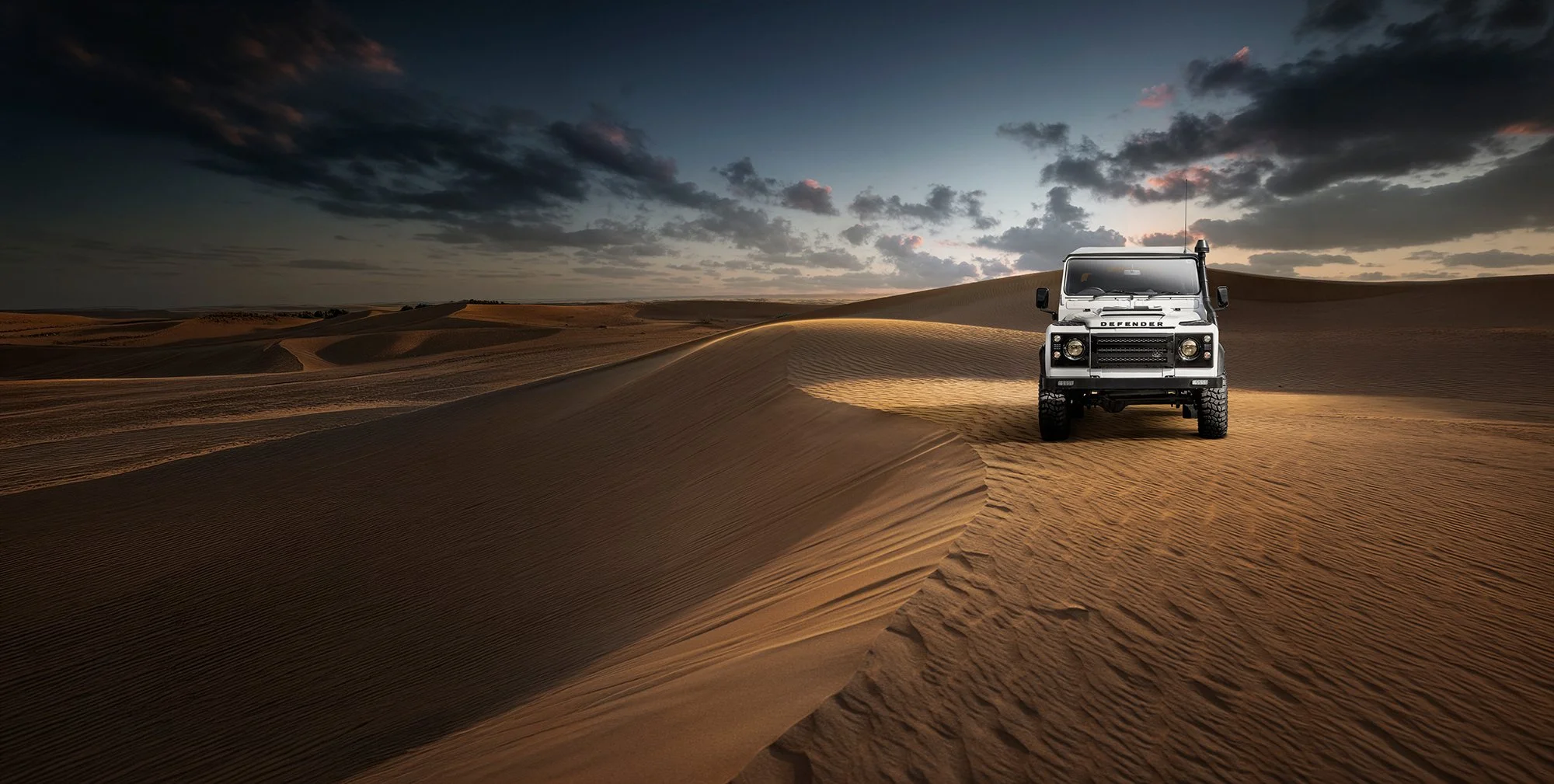 Land Rover Defender in white sat on top of a sand dune in the desert.
