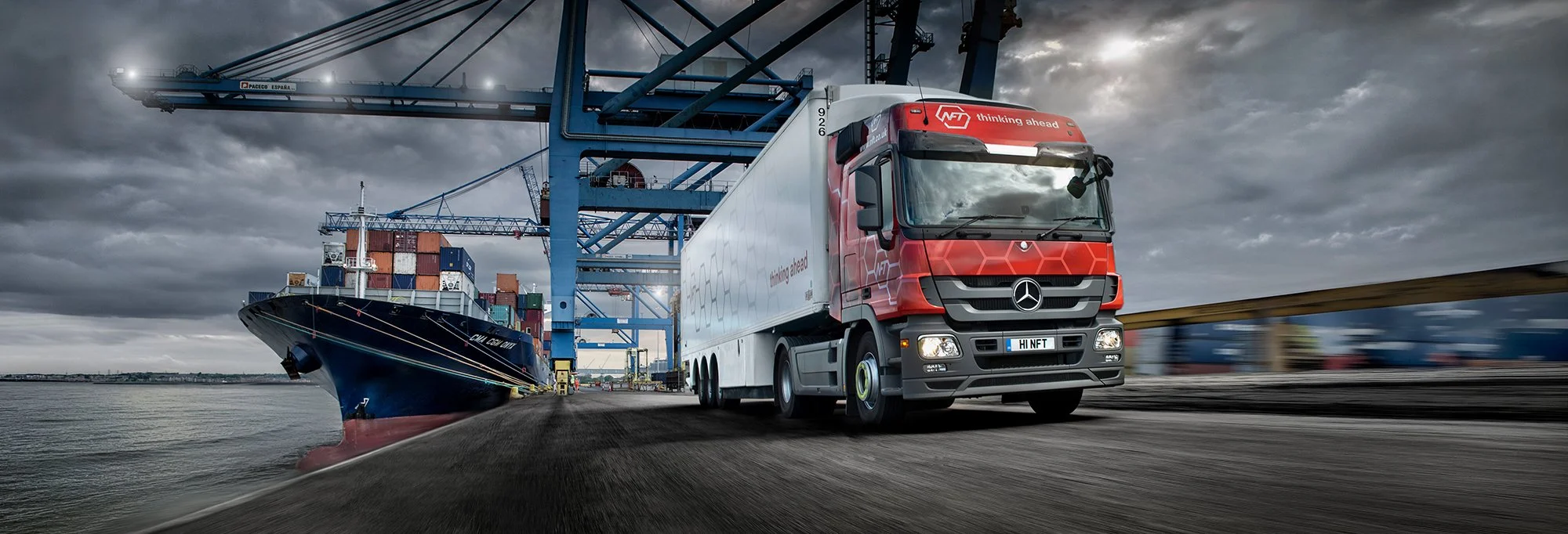 Truck driving on at a port with ship in background.