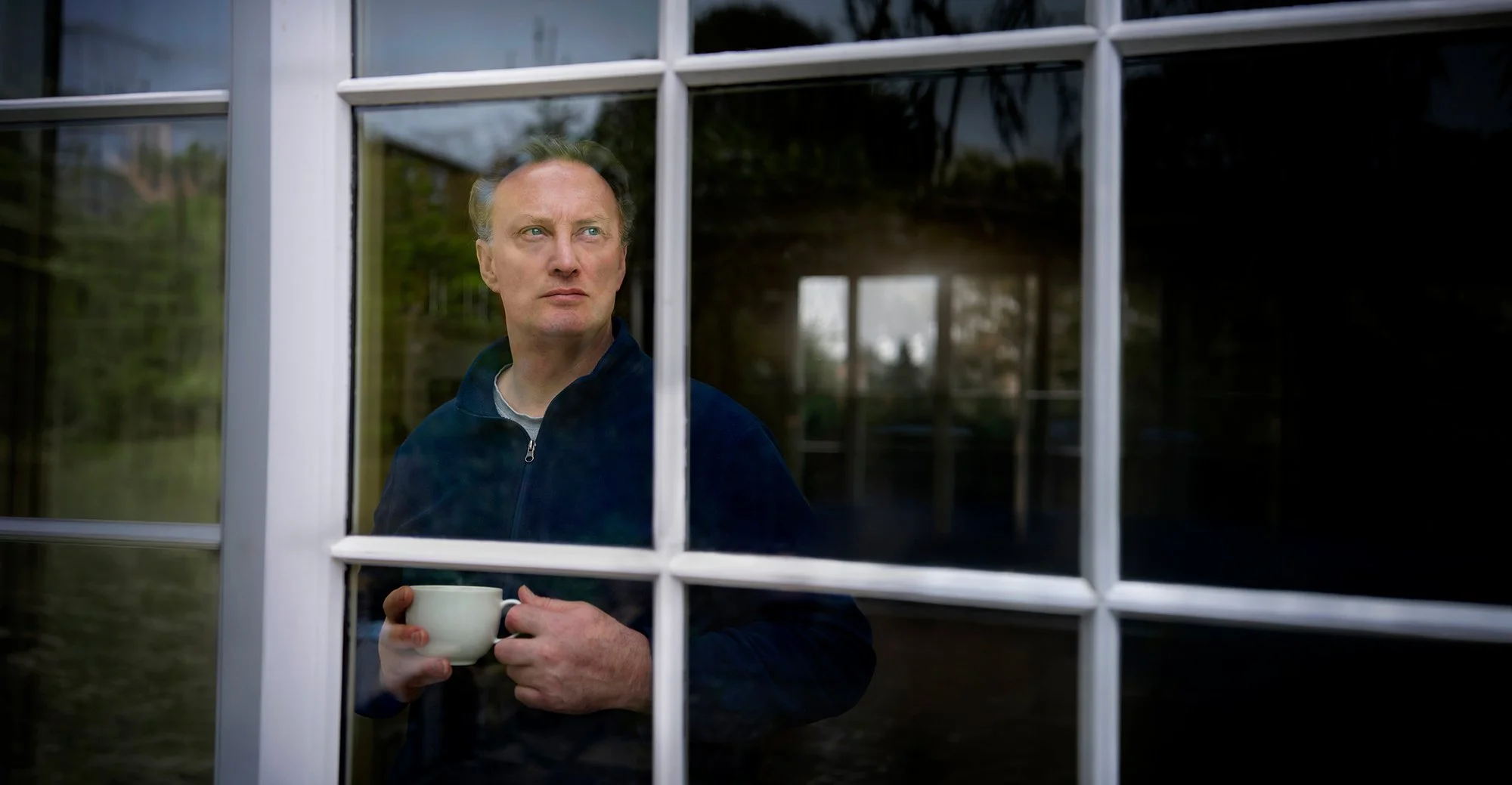 Man looking out of a window with a coffee cup in his hand.