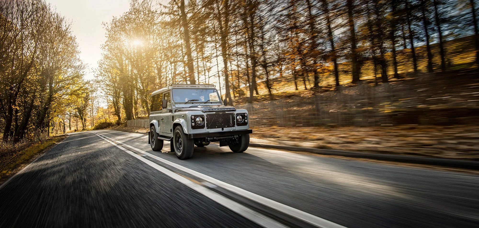 Land Rover Defender driving on a sunny road through a forest.