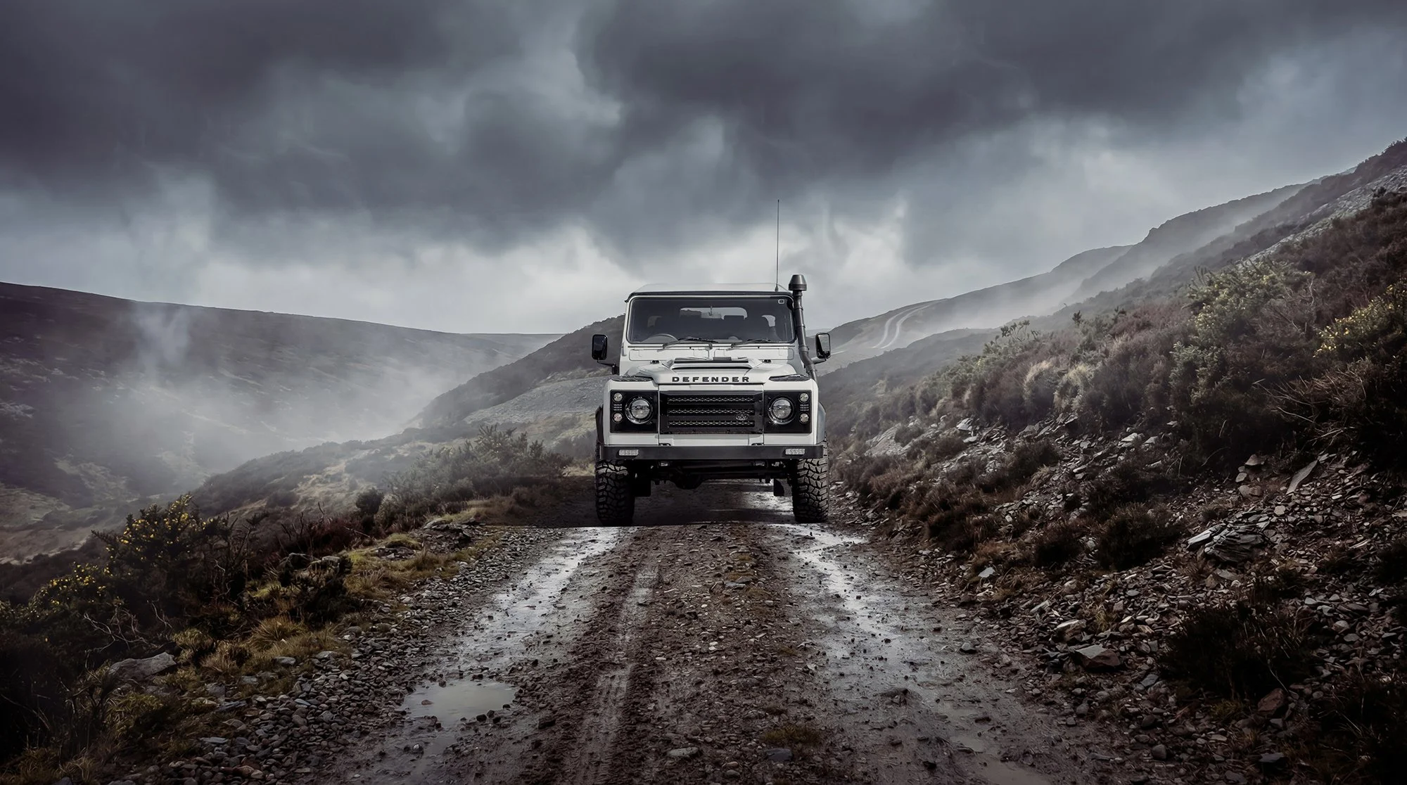White Land Rover Defender on muddy country lane.