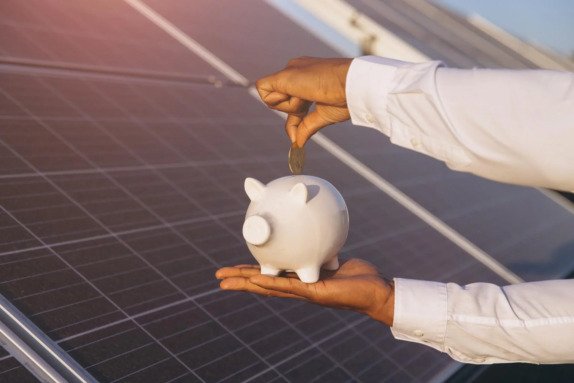 A person in a white shirt holding a white piggy bank while placing a coin into it, with solar panels in the background.