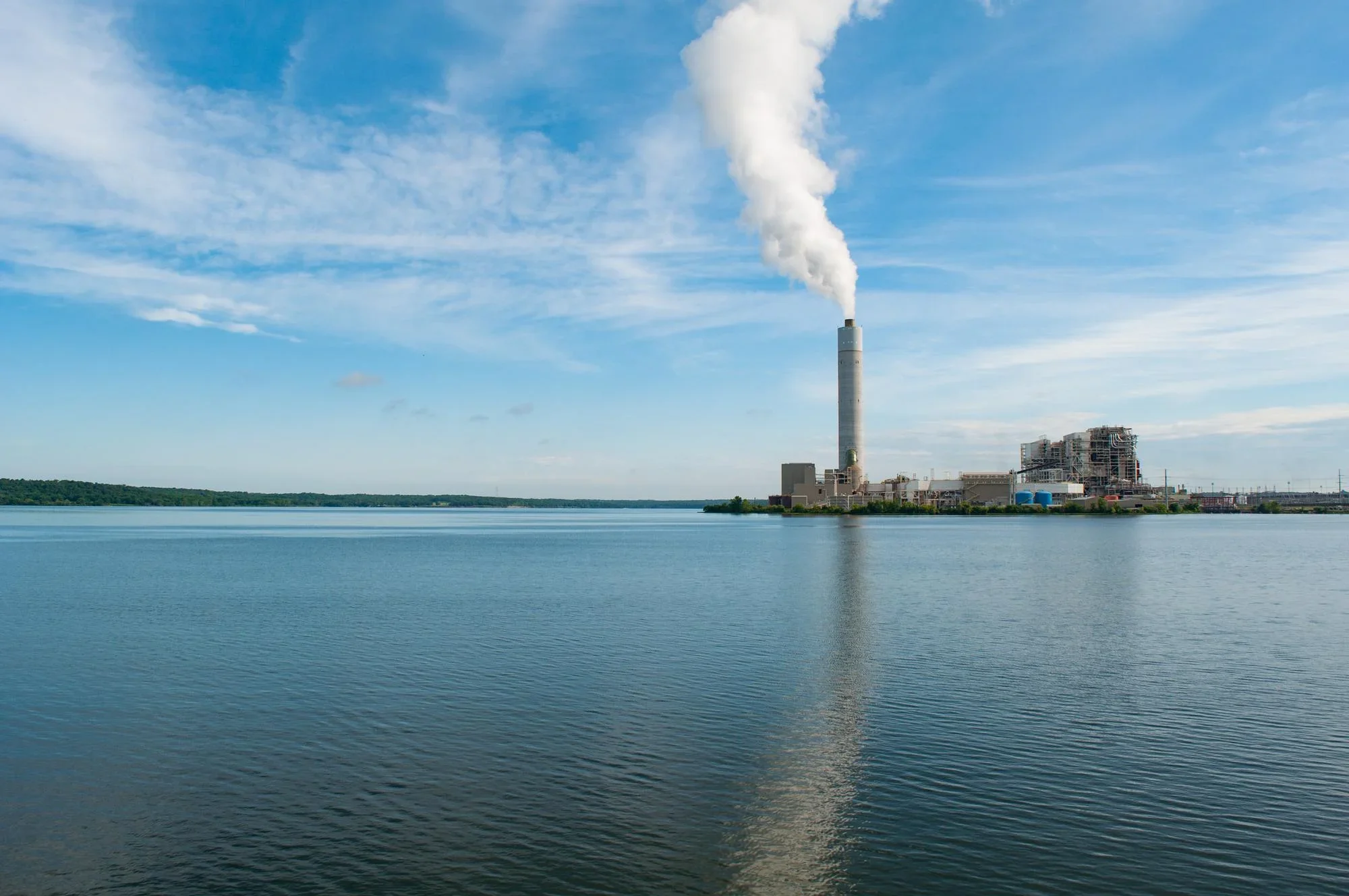A power plant emitting white smoke from a tall chimney near a large body of water with a partly cloudy sky above.