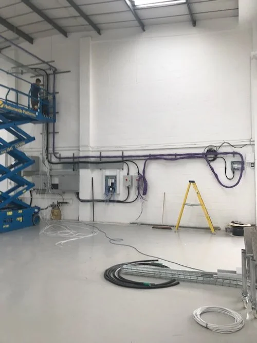 Construction or maintenance area with a blue scissor lift, ladder, cables, and electrical equipment in a large indoor space.