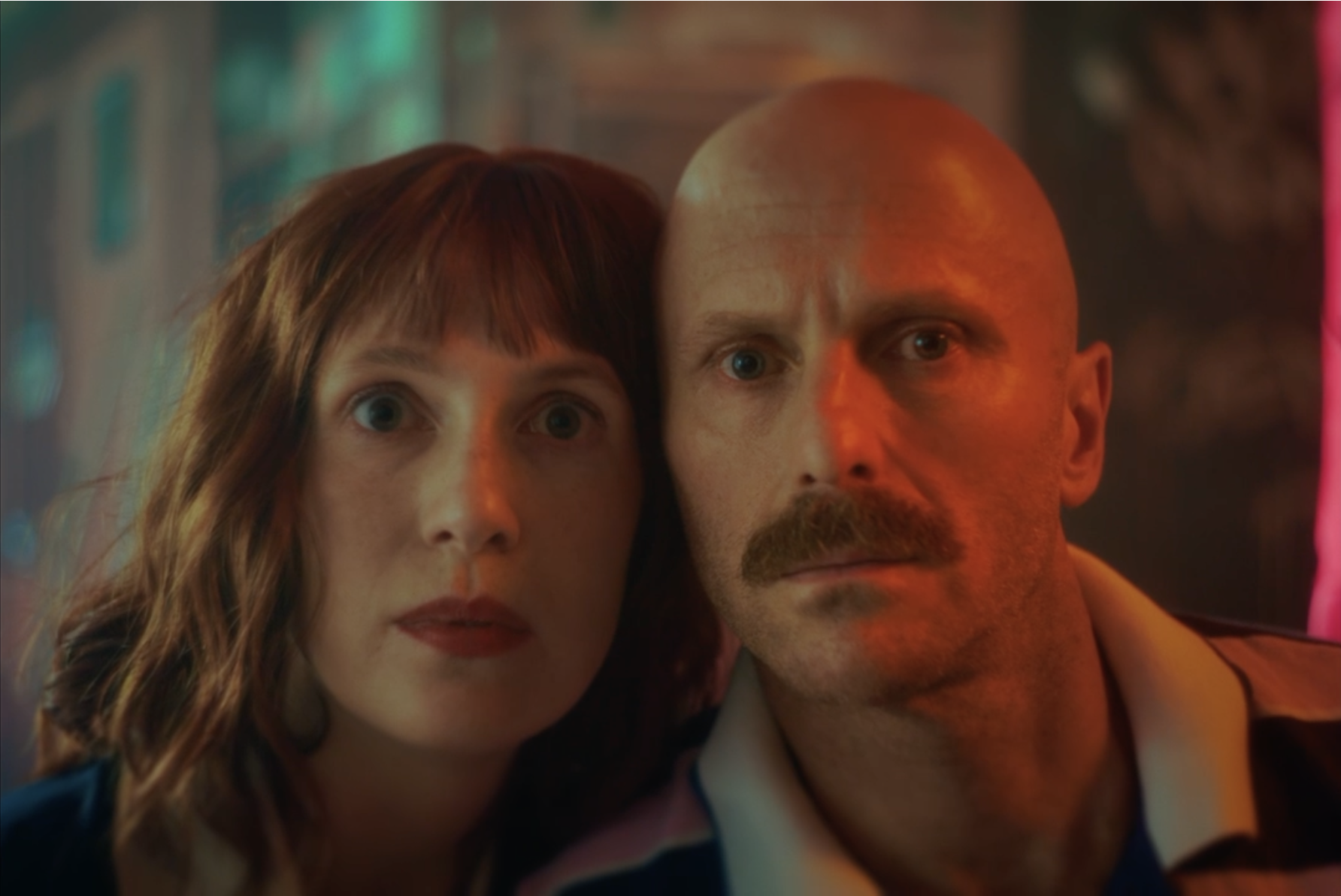 Nahaufnahme von einem Mann und einer Frau, die eng beieinanderstehen, mit einer bunten, neonlichterbeleuchteten Hintergrundszene.