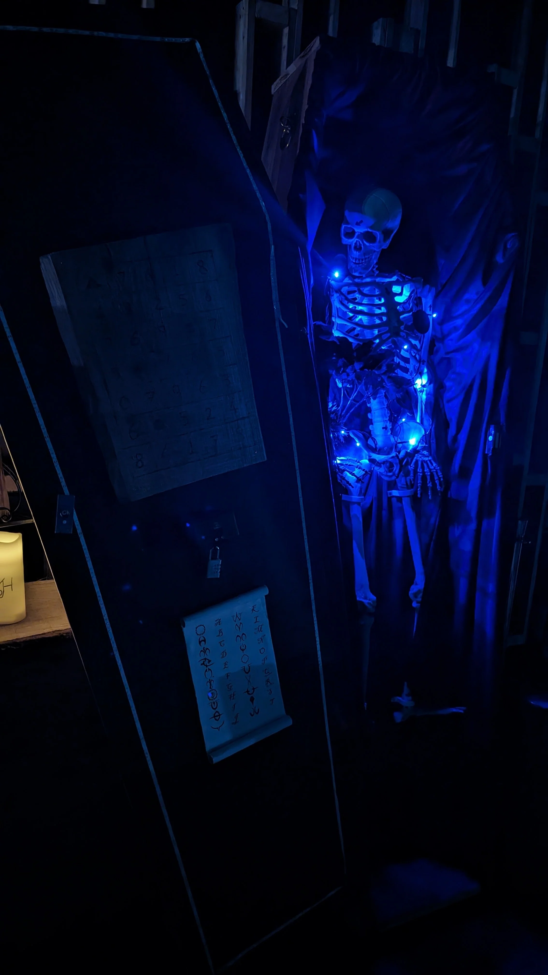 A skeleton decorated with blue string lights standing in a dark room, with a mirror and notes or signs with writings nearby.