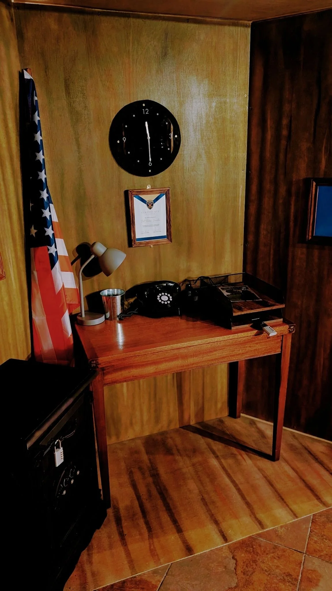 Wood-paneled office corner with a table, vintage rotary phone, desk lamp, and an American flag, with a wall clock and framed certificate on the wall.