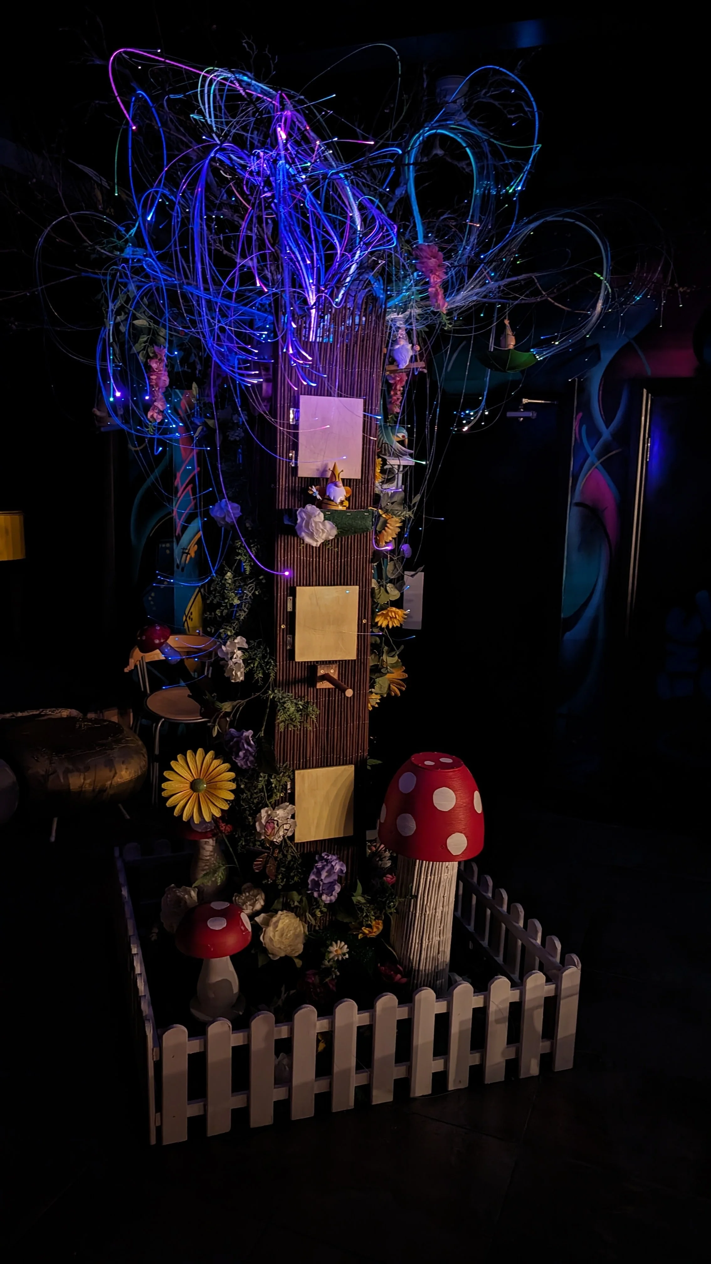 Colorful light display with illuminated wires, artificial flowers, and decorative mushrooms inside a small fenced area.