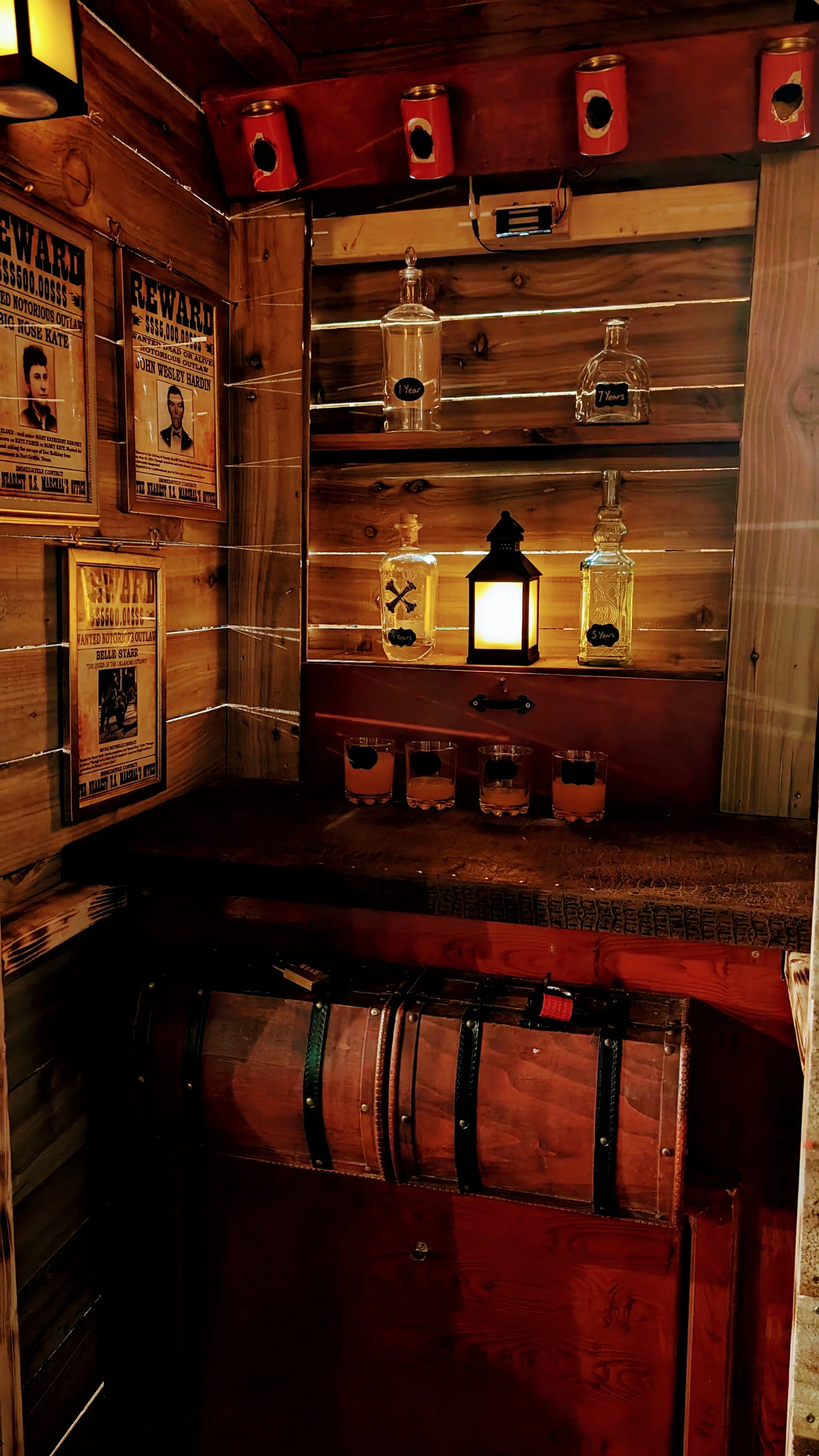 A vintage-style wooden corner with liquor bottles marked with ages, a lantern-shaped light, framed posters of wanted outlaws, and four short glasses filled with pinkish drinks, all in a rustic setting.