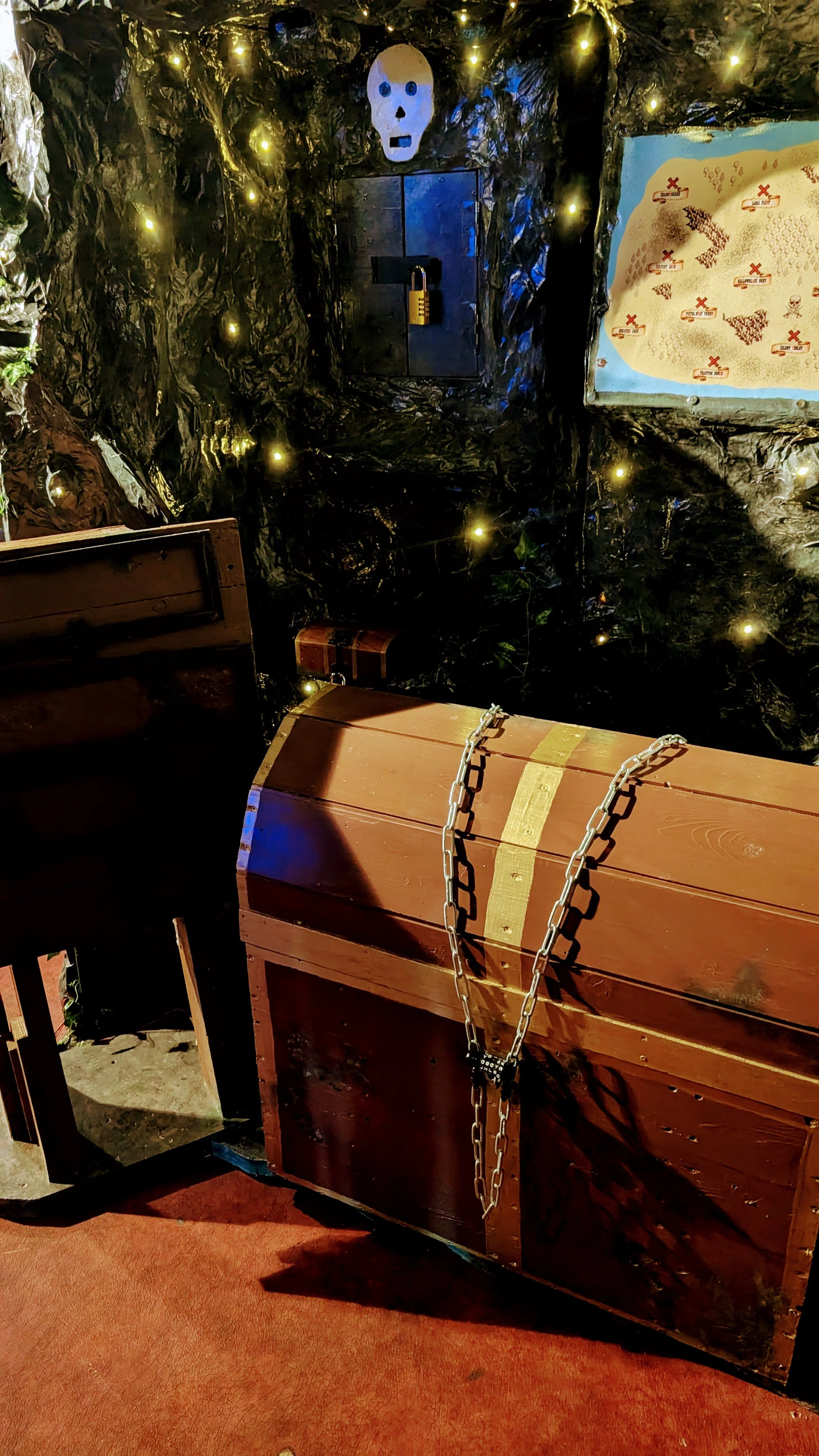 A wooden treasure chest with a metal chain lock sits on a red carpet. Behind it is a dark, cave-like backdrop with a lit skull decoration and a map with red markings and cross symbols, illuminating a spooky scene.