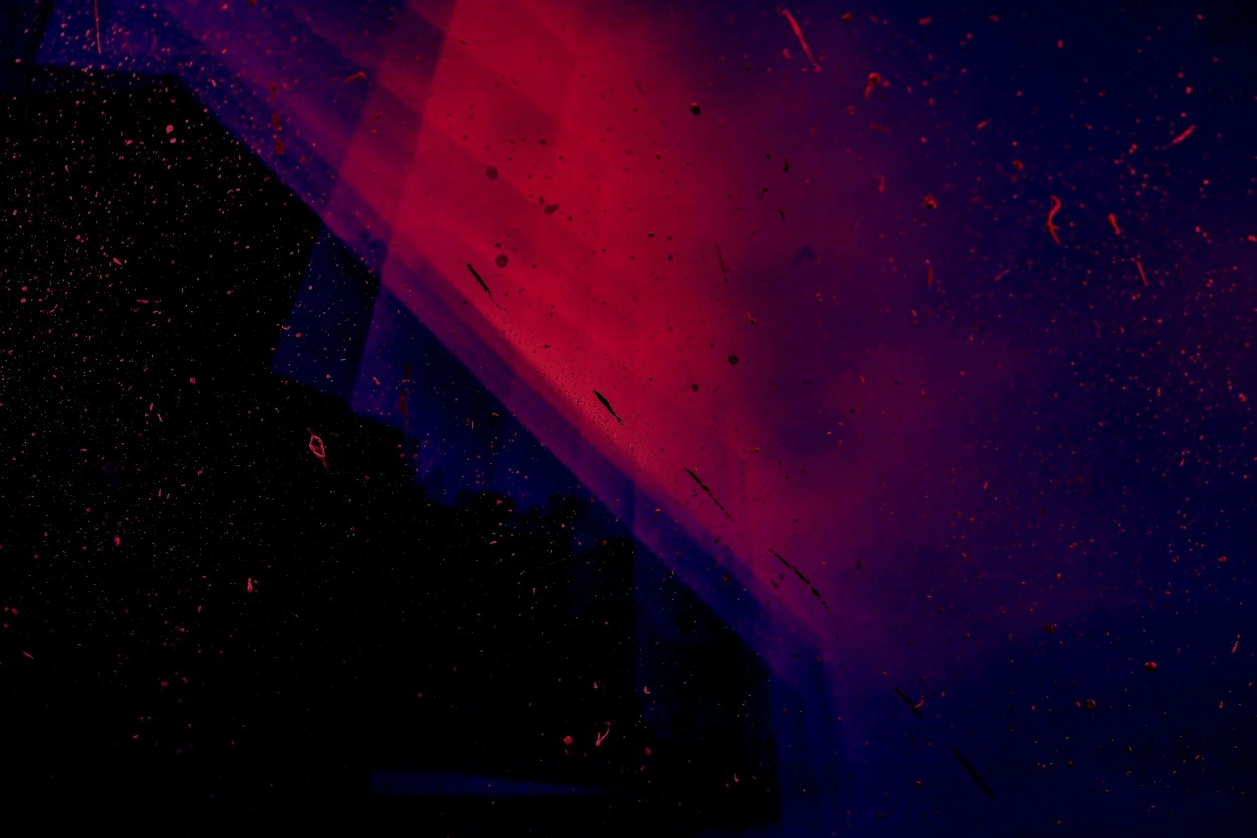 Abstract image with dark background, featuring a large red and purple gradient shape, accompanied by scattered small particles and splashes, creating a cosmic or space-like effect.