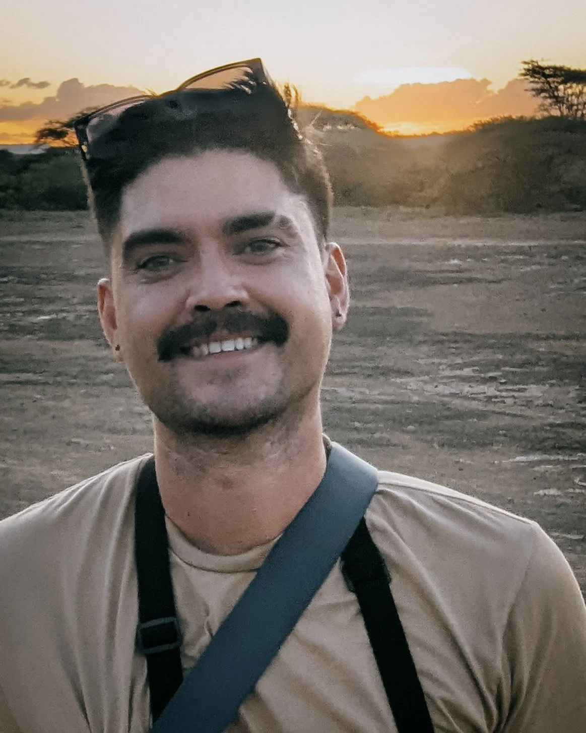 A smiling man with dark hair, a mustache, earring, and sunglasses on his head, wearing a beige shirt and a camera strap, standing outdoors during sunset with a landscape of dry terrain and sparse trees in the background.