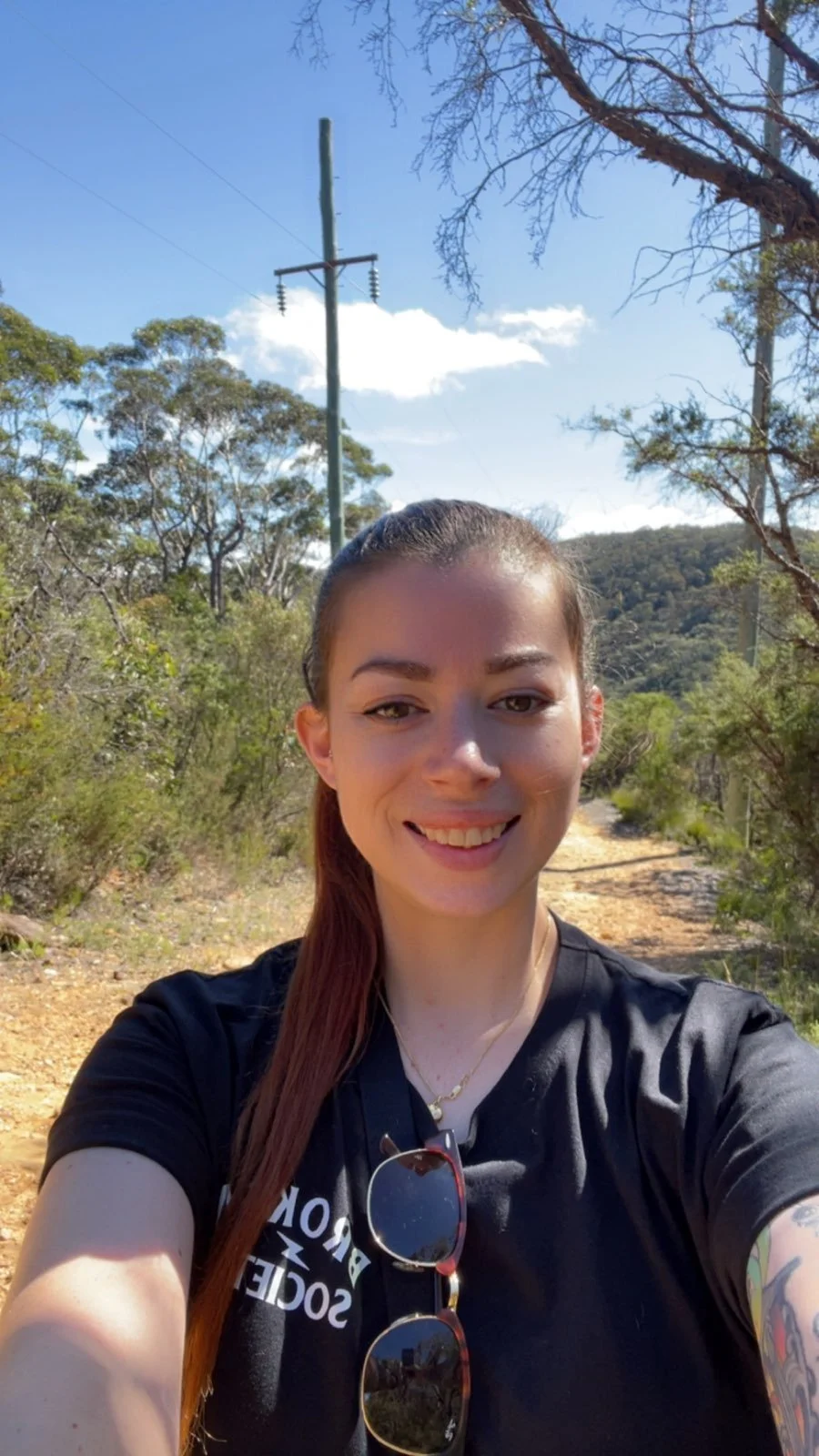 A woman taking a selfie outdoors on a sunny day with trees, hills, and a utility pole with power lines in the background.