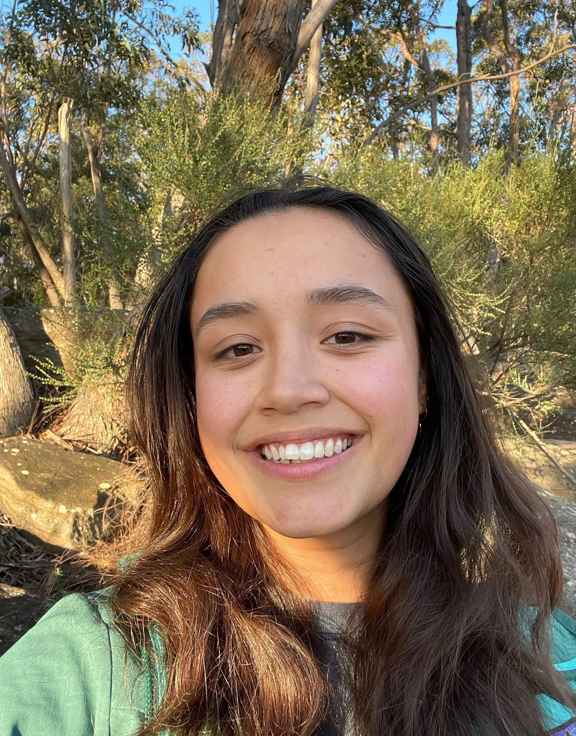 A young woman with long dark hair, smiling, outdoors in front of trees with sunlight illuminating her face.
