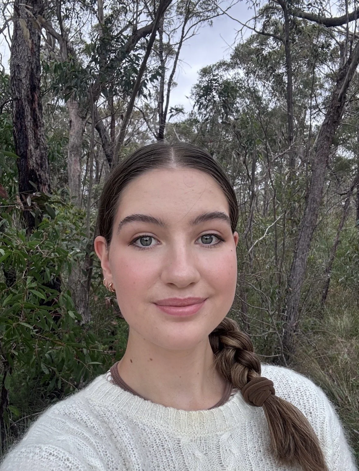 A young woman with braided hair and hoop earrings standing outdoors among trees and shrubs, wearing a cream-colored sweater.