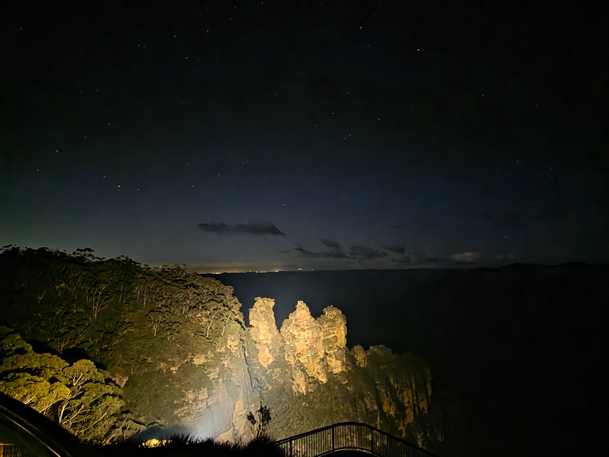 Nighttime view of rugged coastal cliffs with greenery, illuminated from below, under a starry sky with clouds and a faint horizon line.