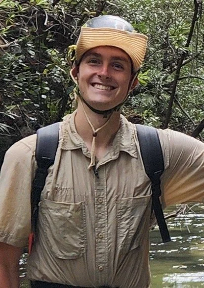 A smiling man wearing a helmet, khaki shirt, and backpack in a wooded outdoor setting.