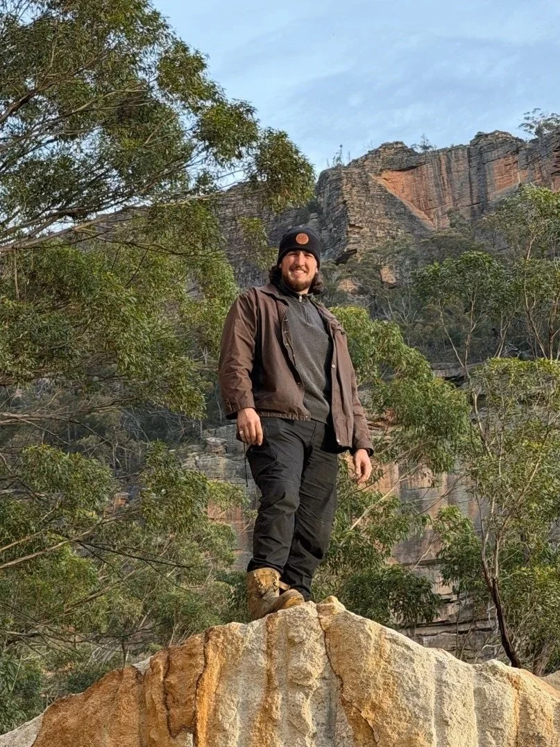 A man standing on a large rock in a natural outdoor setting with trees and cliffs in the background.