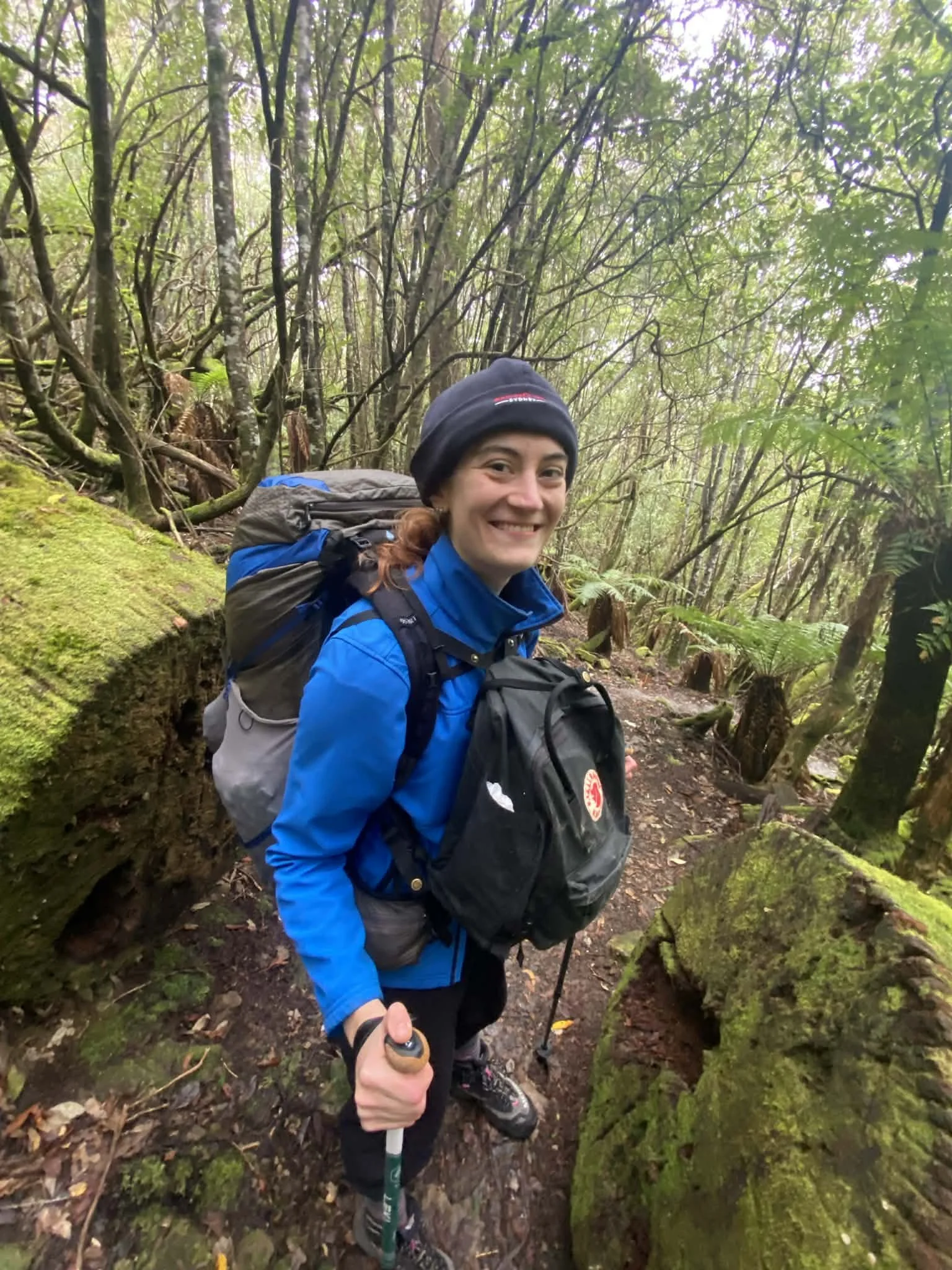 A woman hiking in a forest, smiling, wearing a blue jacket, black beanie, and carrying a backpack. She's holding a hiking pole and surrounded by lush green trees and moss-covered rocks.
