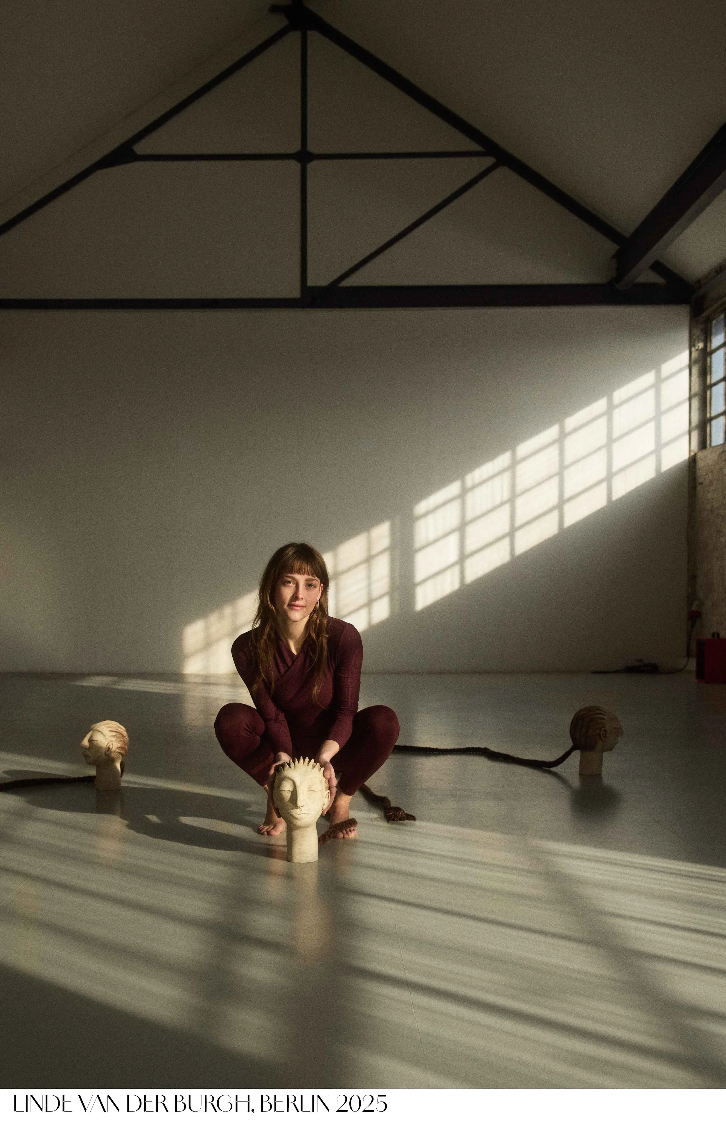 Analog portrait of a performance artist Linde van der Burgh with sculptural heads arranged around her. Daylight casts a window-shaped pattern onto the wall behind her.  Tereza Konarikova photography