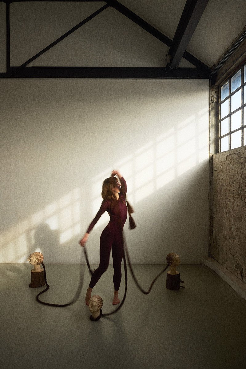 Performance artist Linde van der Burgh in a dark red overall moves around sculptural headpieces during the Womb-hood performance at Chaos Berlin. Sunlight creates a window-shaped shadow on the wall behind her as she moves through the space.