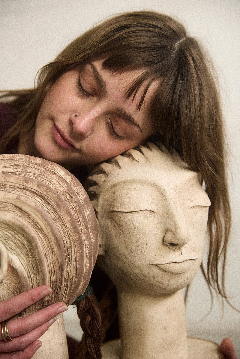 Calm portrait of a young artist Linde van der Burgh eyes closed holding her sculptural heads, photographed in diffused natural light by Tereza Konarikova