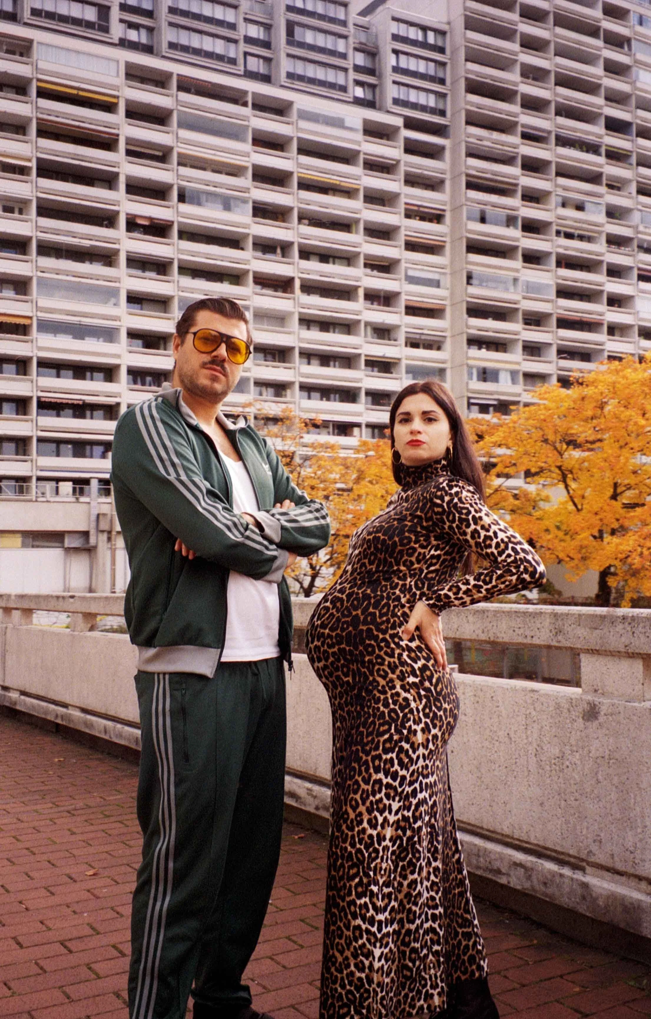 Editorial pregnancy portrait of a couple in Munich, with a pregnant woman in a leopard dress and her husband in an Adidas tracksuit, photographed against Plattenbau architecture of Olympiadorf münchen.