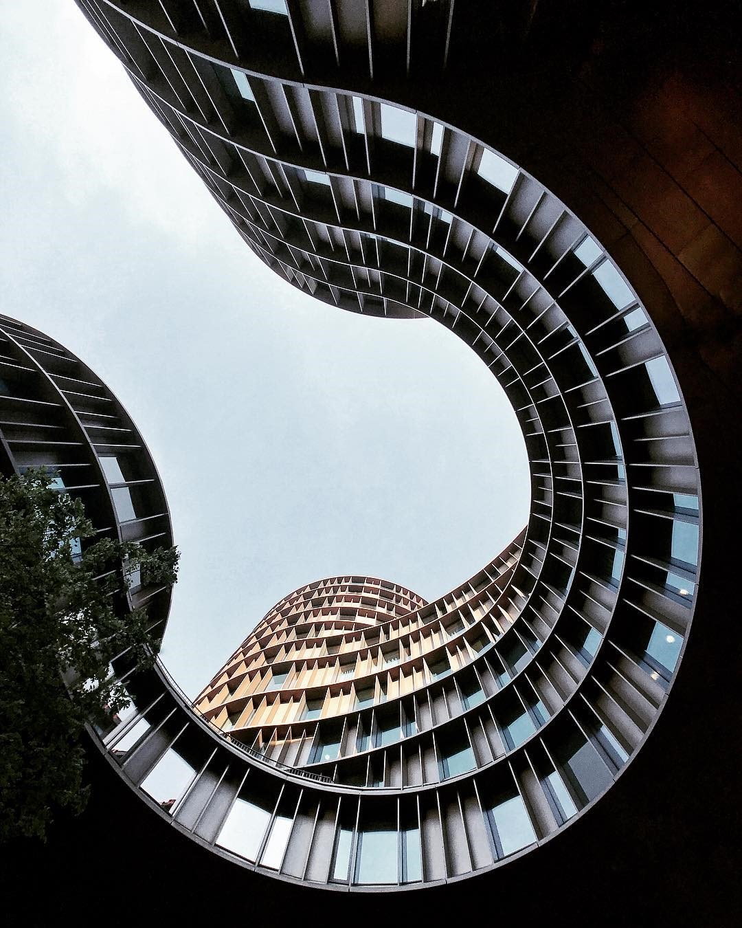 Looking up at a modern, spiral-shaped multi-story building with curved balconies and extensive glass windows, against a cloudy sky.
