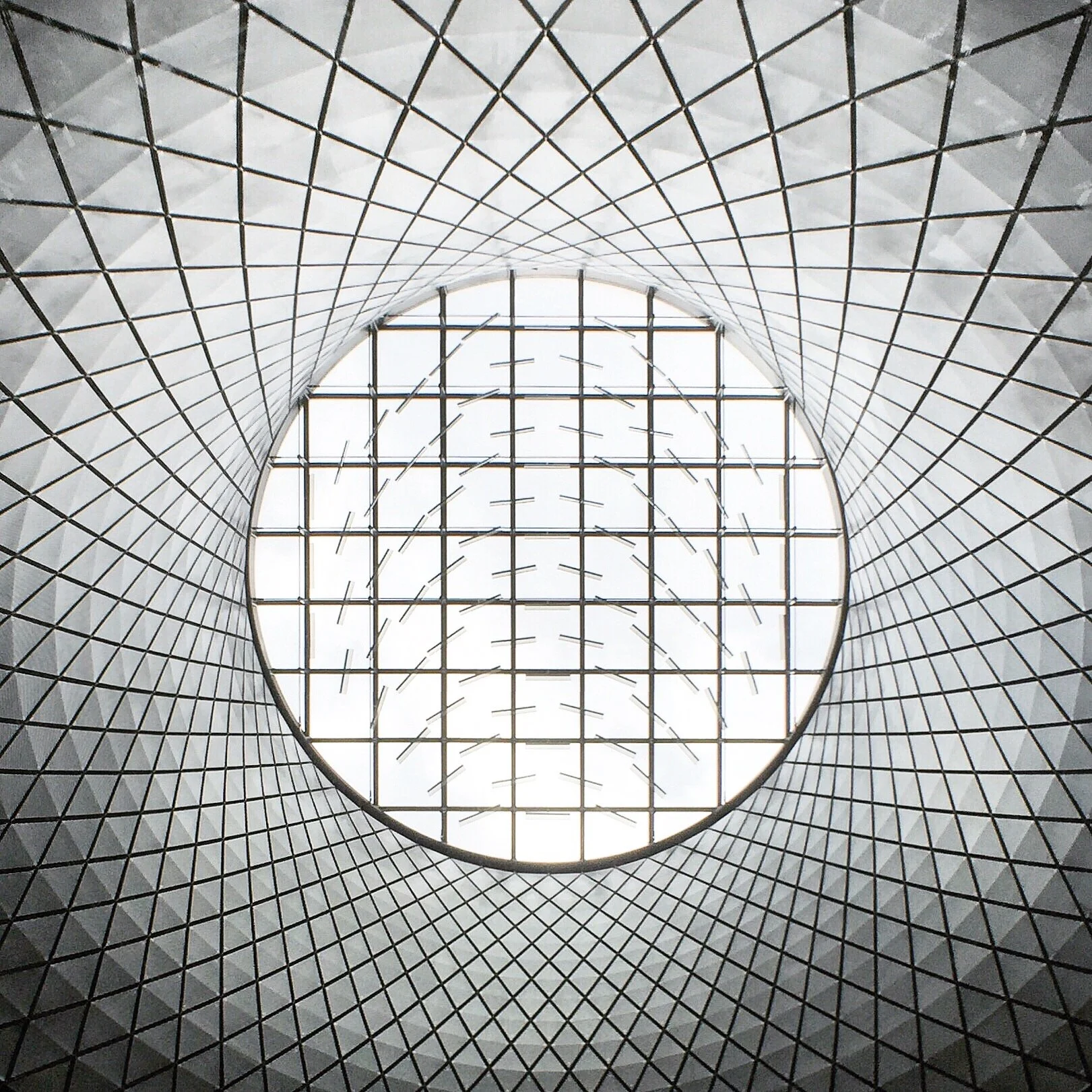 Inside view looking up at a modern, geometric ceiling with a large central skylight. The architecture has a grid pattern with a circular opening at the center, allowing natural light to flood the space.