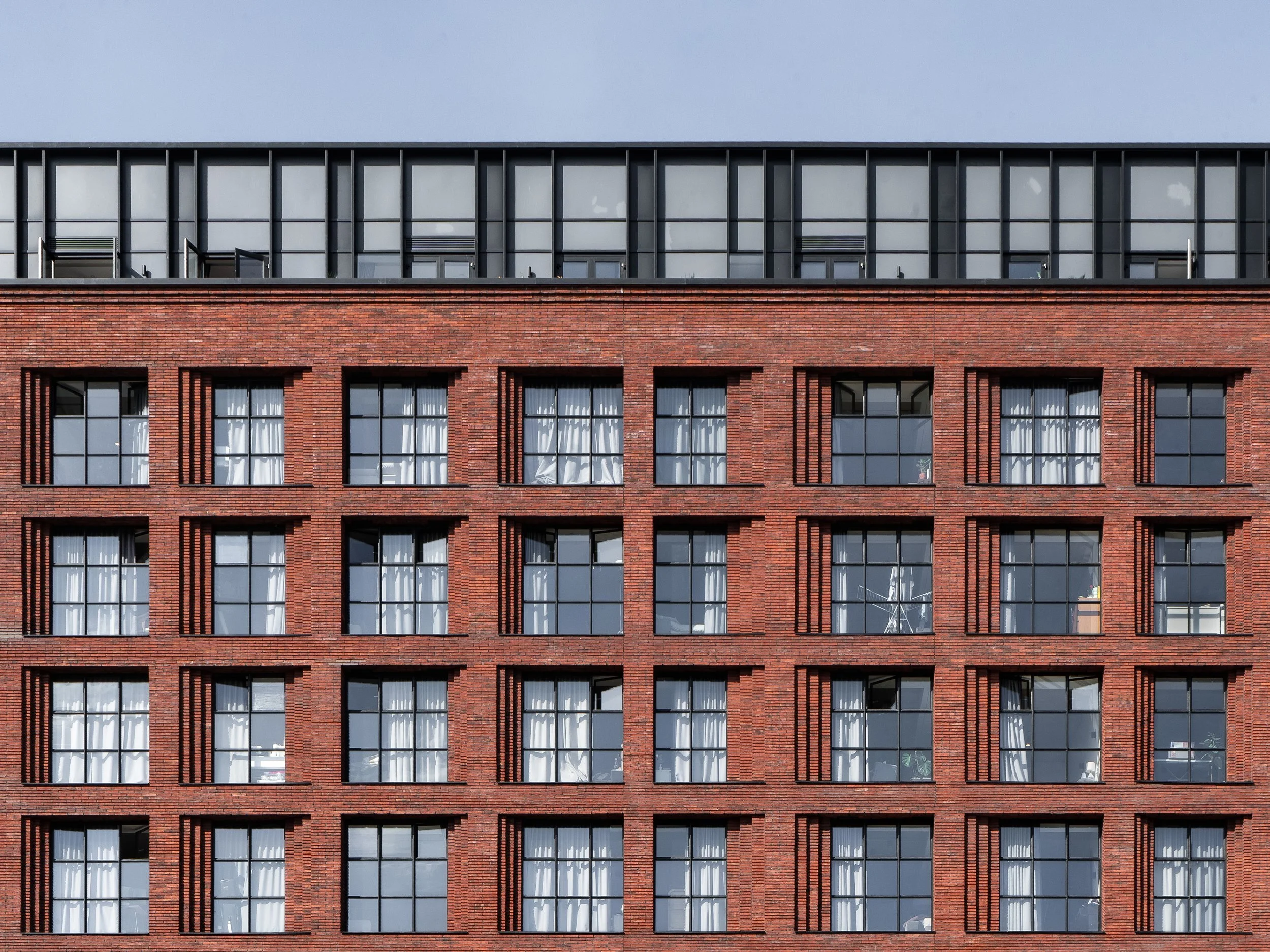 A multi-story brick building with large windows and open window shades, with a section of modern black metal and glass on the top floor.