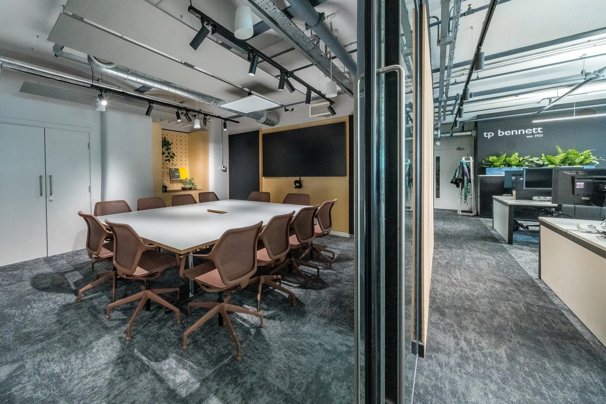 Modern conference room with a large table and ergonomic chairs, glass wall, and visible office equipment, with a logo reading 'tp bennett est. 1921' on the wall. Manchester