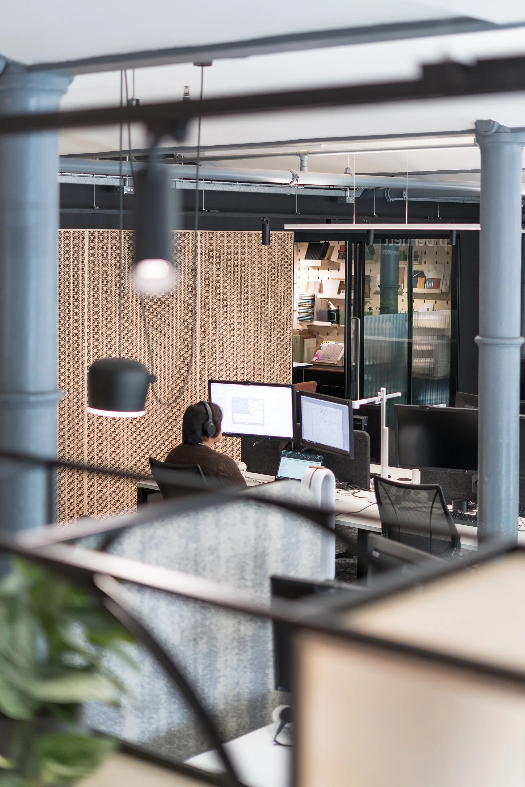 Office workspace seen through a black metal railing, with multiple computer monitors, a person wearing headphones working at a desk, and a wooden wall with bookshelves in the background.