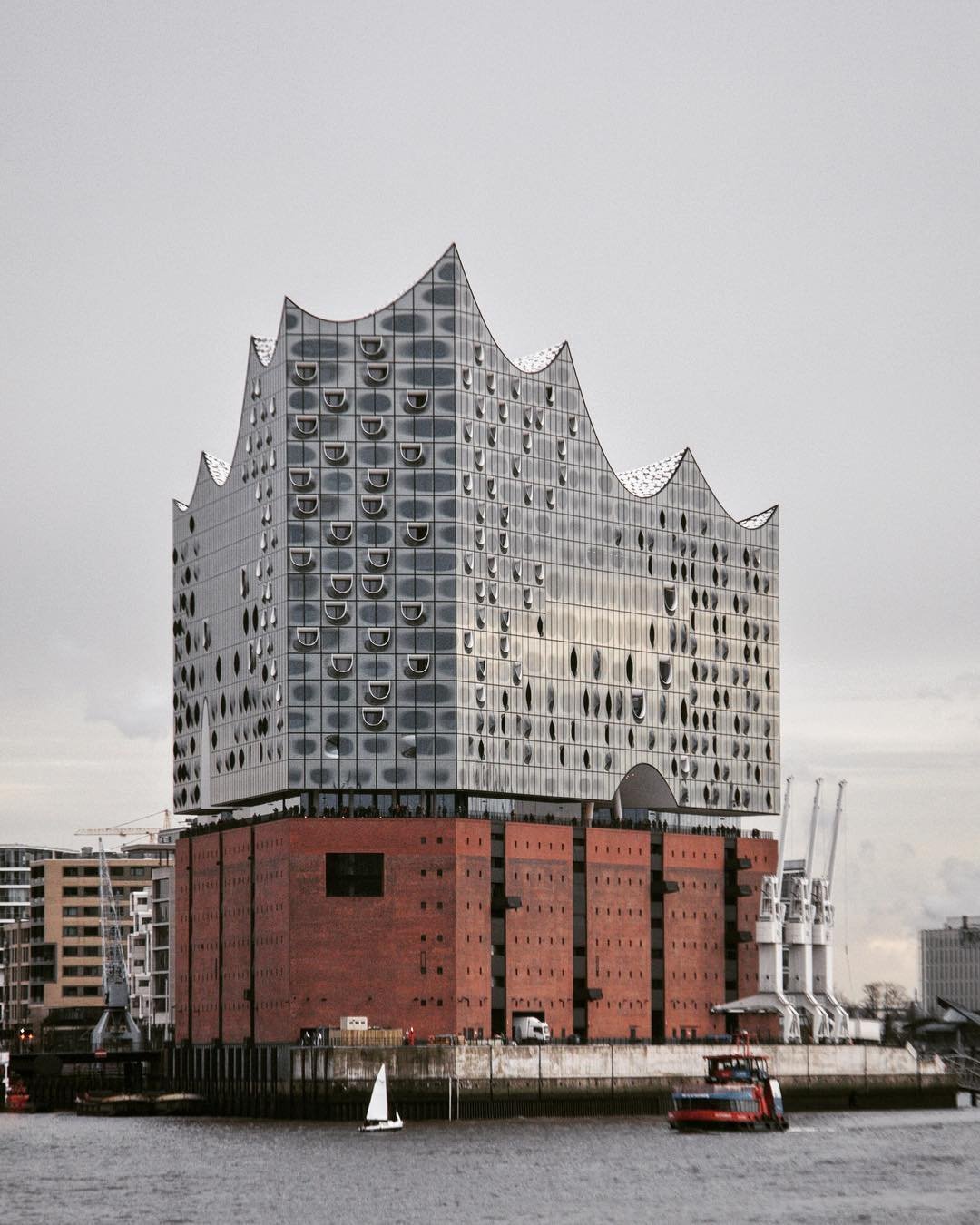 Modern building with reflective glass and unique, jagged roof design, situated along a waterway with boats and surrounding cityscape.