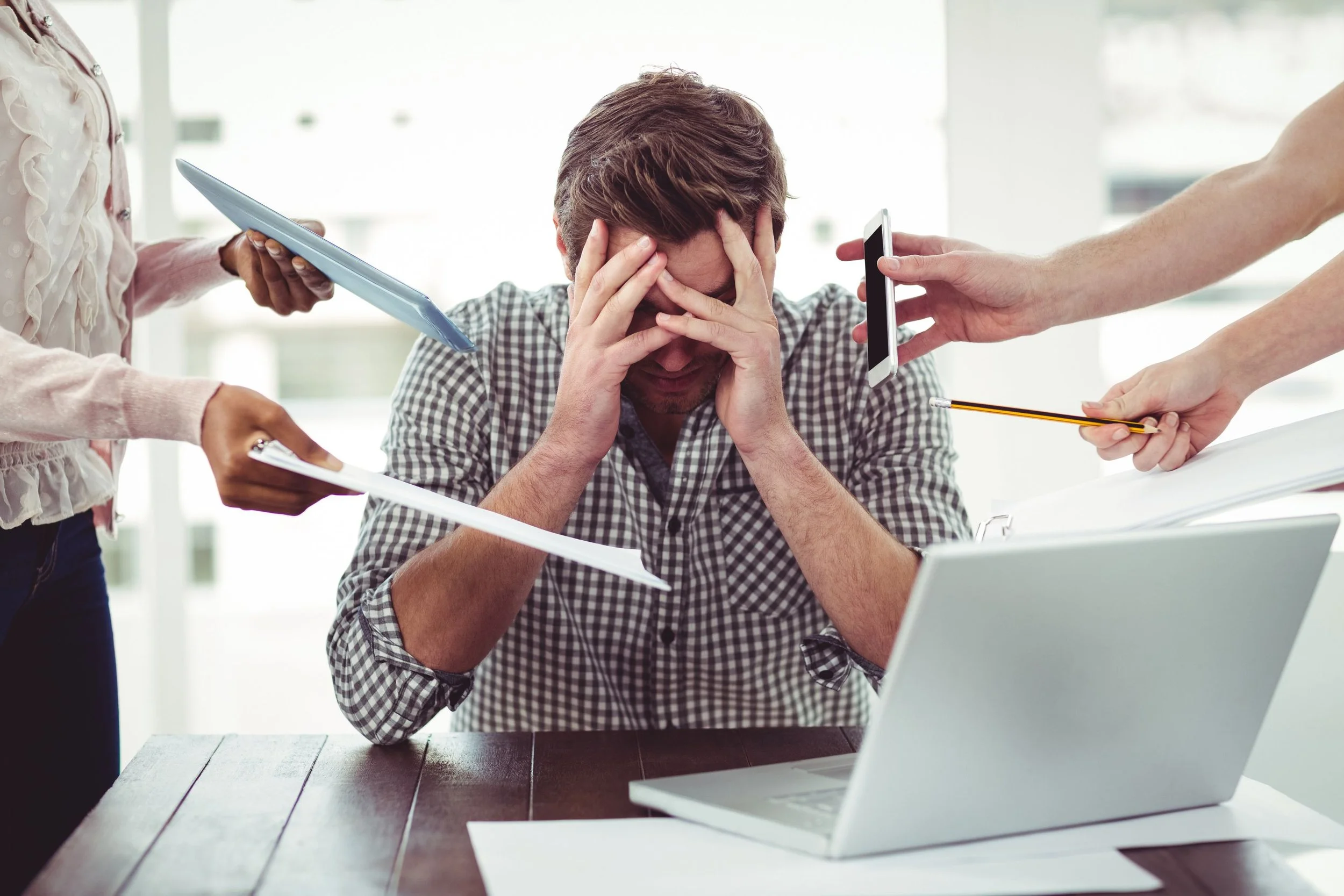 A man sitting at a desk with his hands on his head, appearing stressed or overwhelmed as multiple hands reach out to him with documents, a phone, a tablet, and a pen.