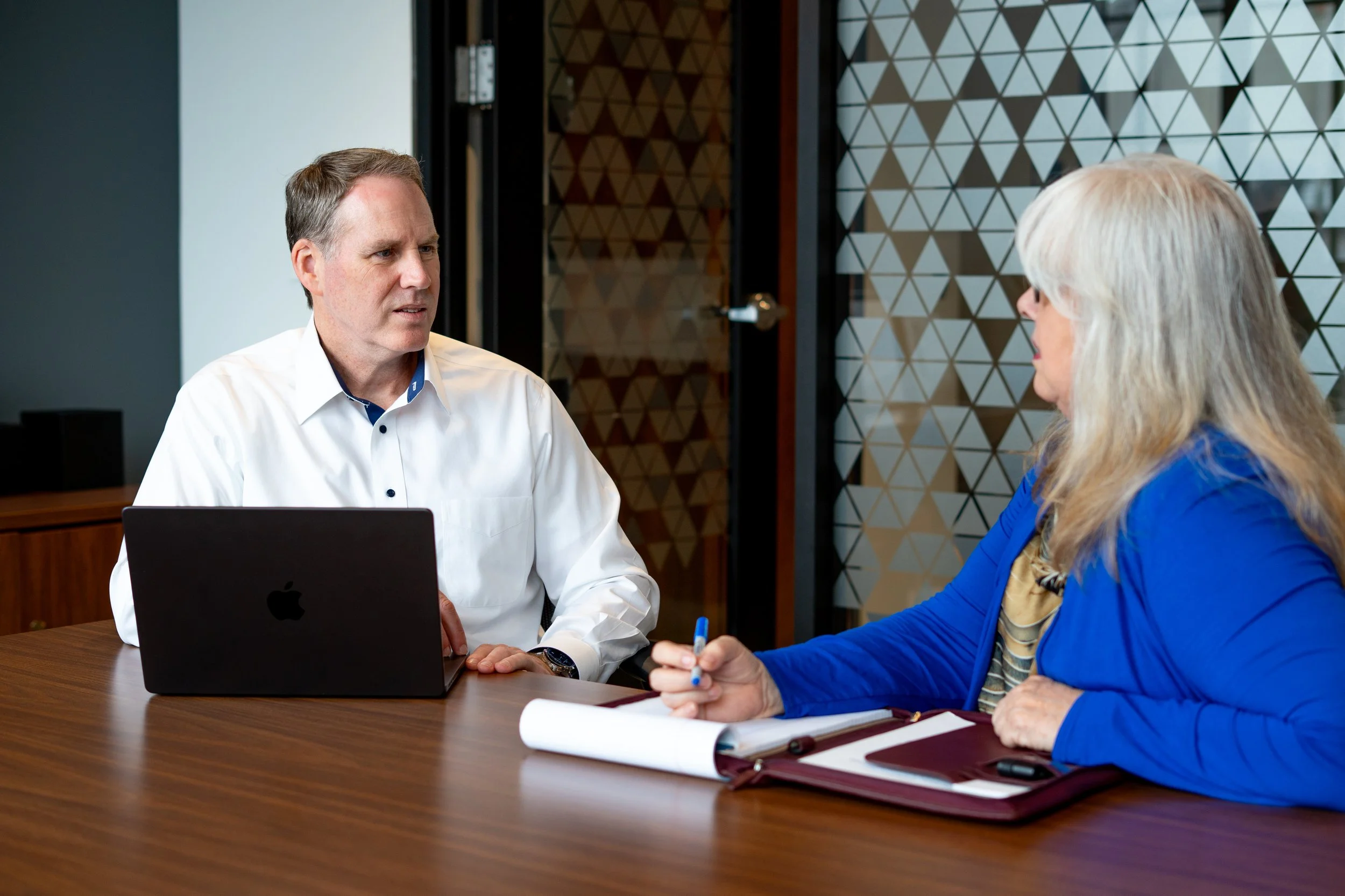 A man and woman having a business meeting in a conference room with a wood table and geometric patterned wall.