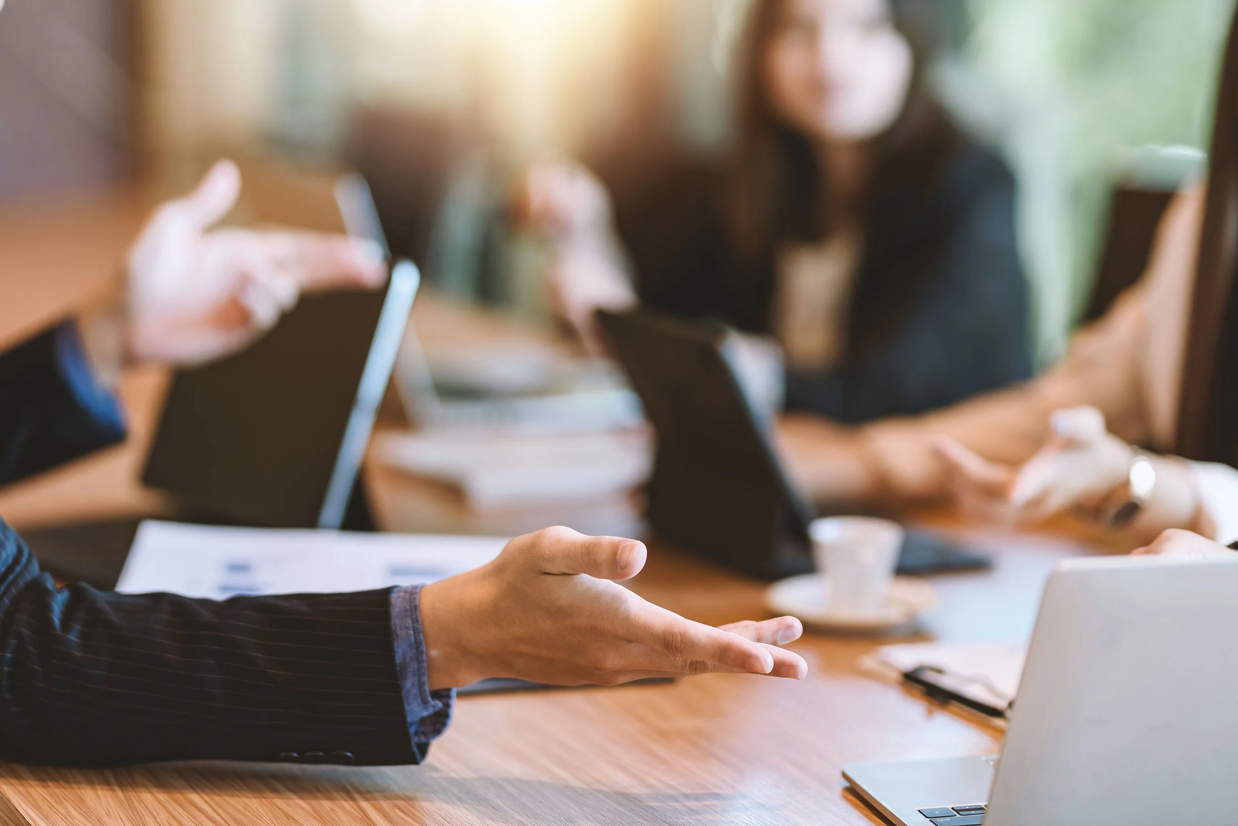 Man asks question to a group of business professionals sitting at a wood table with their laptops open