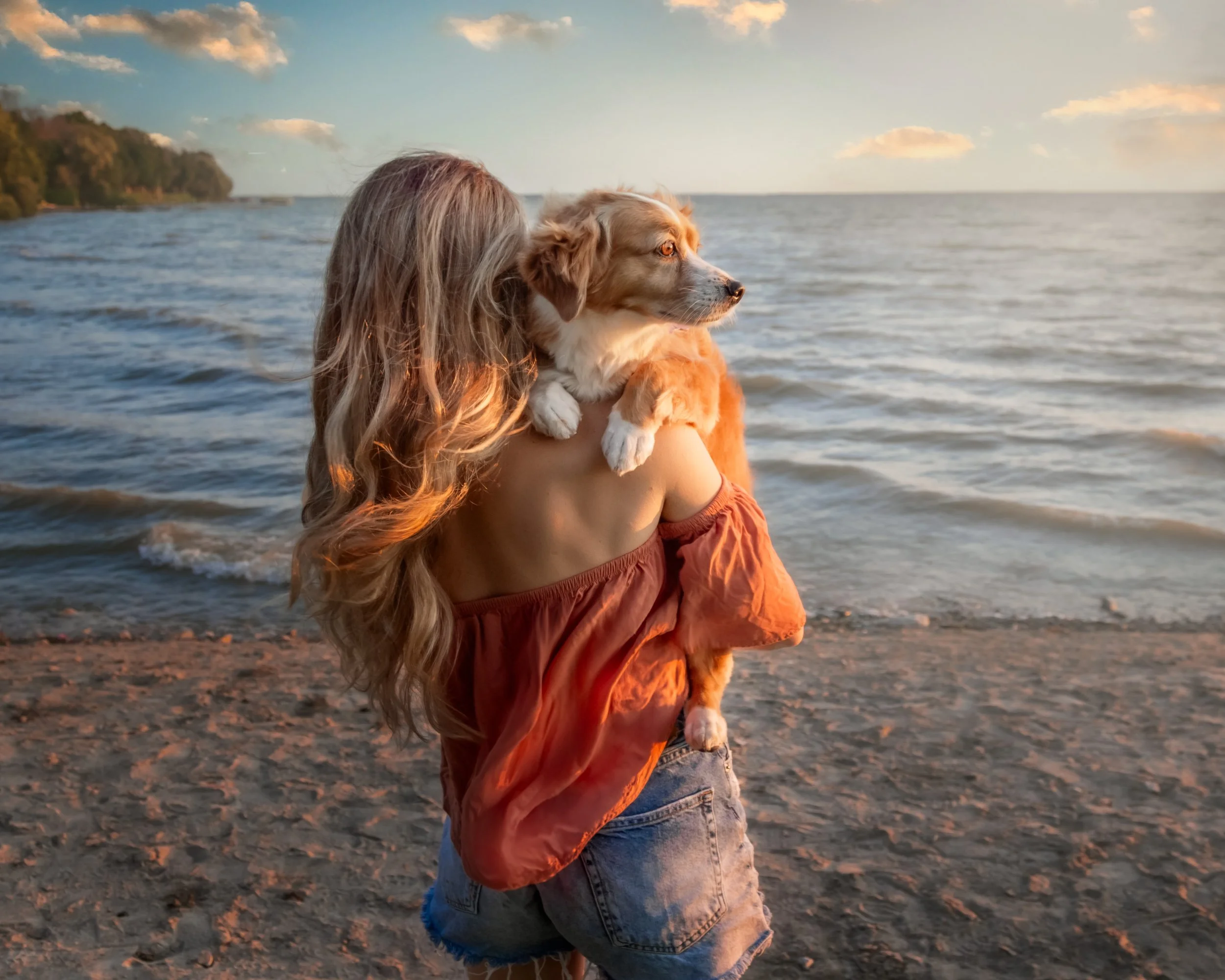 A woman with long hair in a red off-the-shoulder top and denim shorts holding a dog on a beach at sunset, with the ocean and sky in the background.