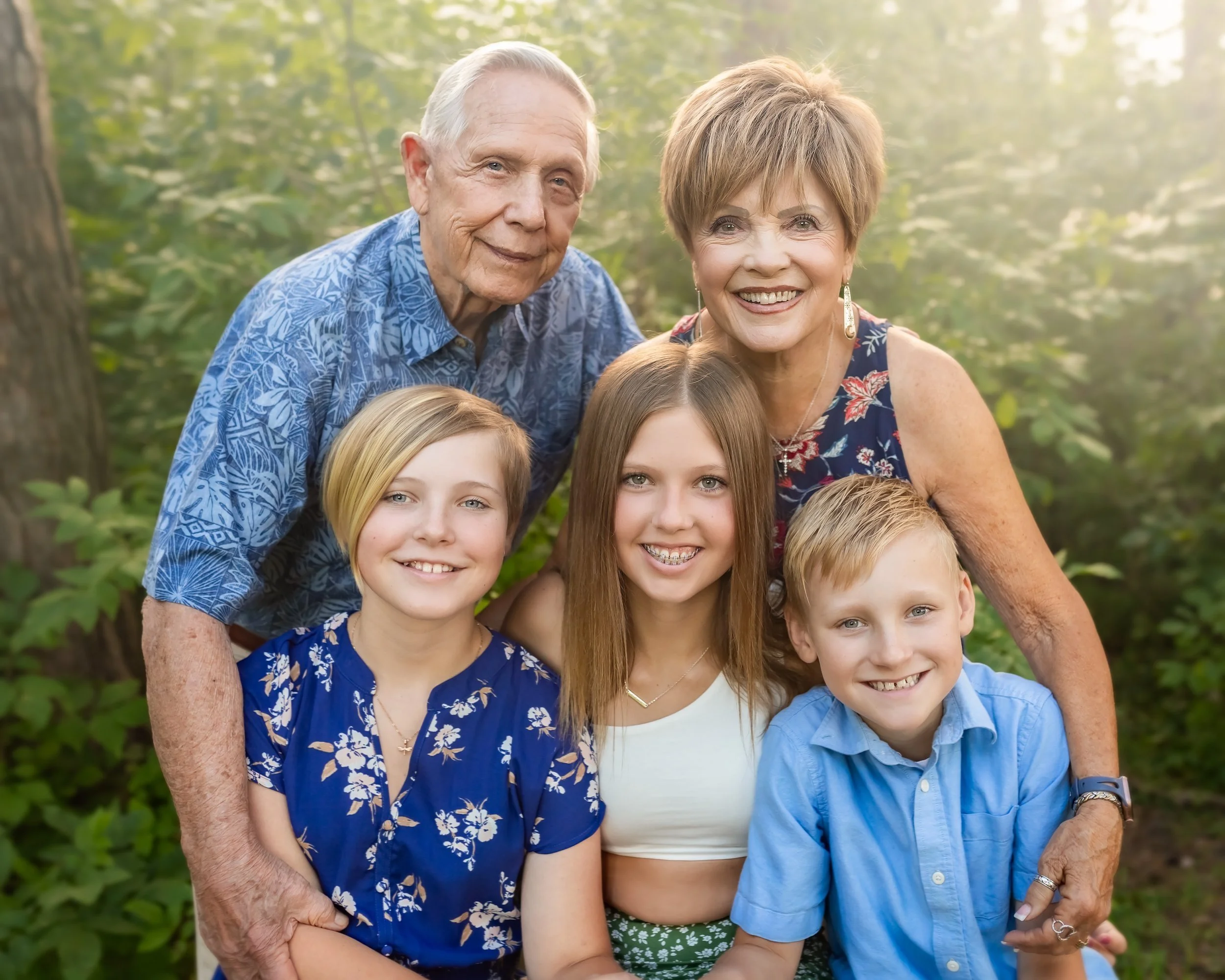 Family of six, including grandparents, mother, father, and three children, smiling outdoors in a wooded area during daytime.