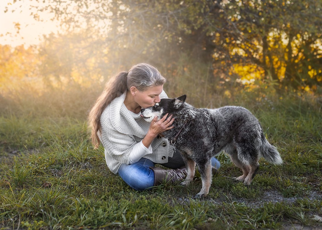 A woman in a white sweater sitting on the grass holds and kisses a black and white dog with a forest background during autumn.