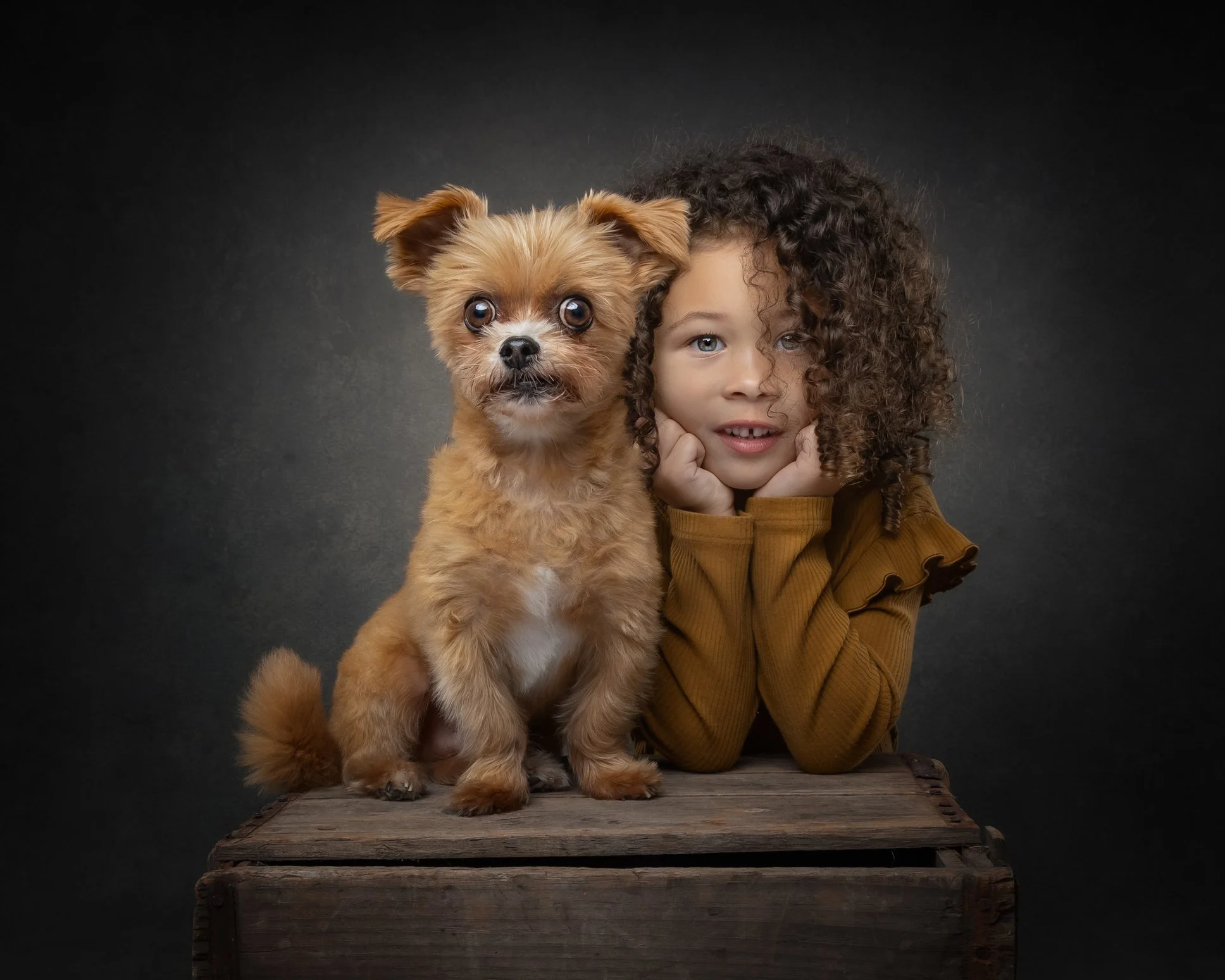 A young girl with curly hair and blue eyes, resting her chin on her hands, next to a small, fluffy dog with large eyes and perky ears, on a wooden surface against a dark background.