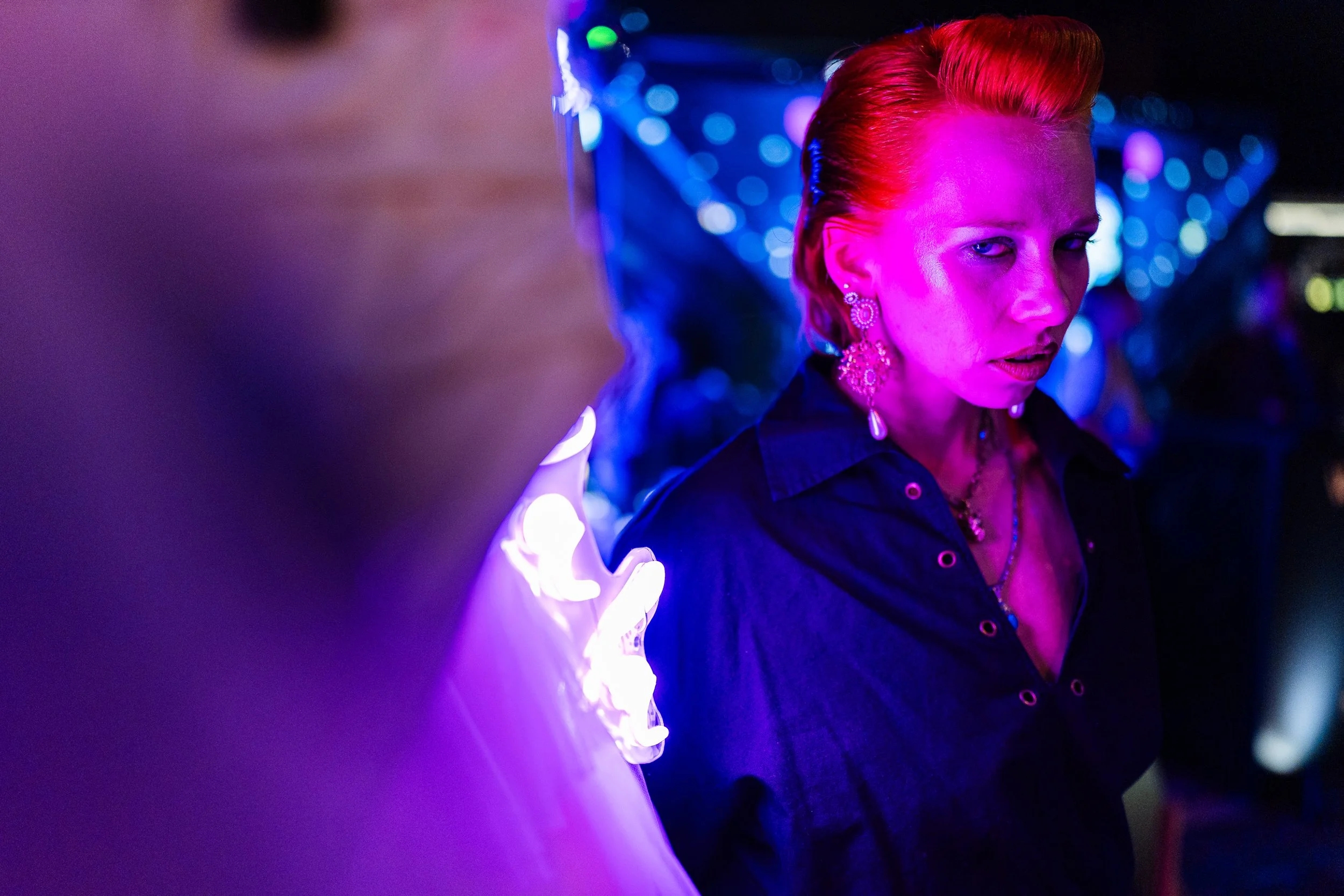 A woman with red hair and earrings looking at a neon-lit sign or object in a dark setting with colorful bokeh lights in the background.