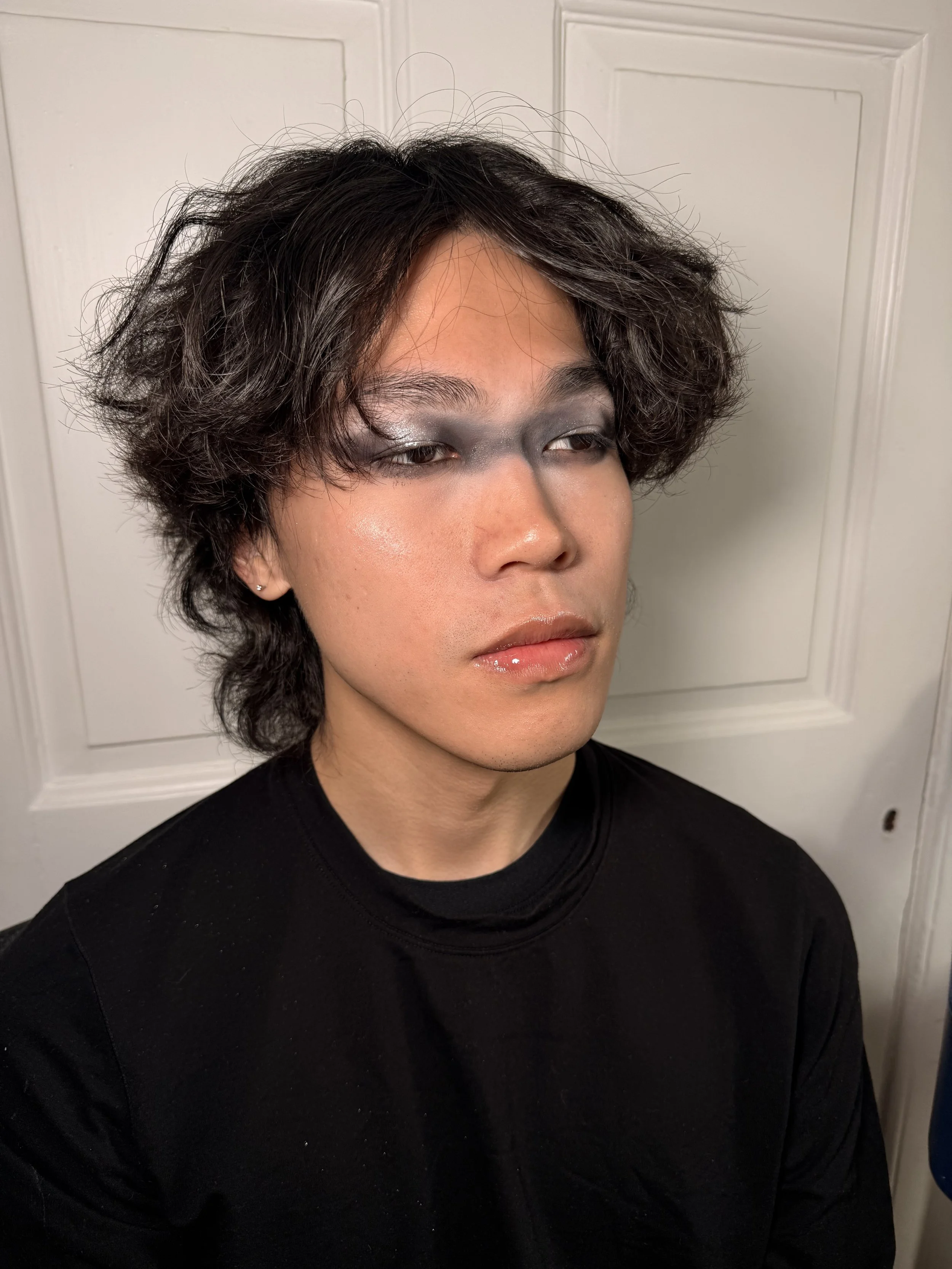 A person with short, curly black hair and makeup, wearing a black shirt, standing in front of a white door.