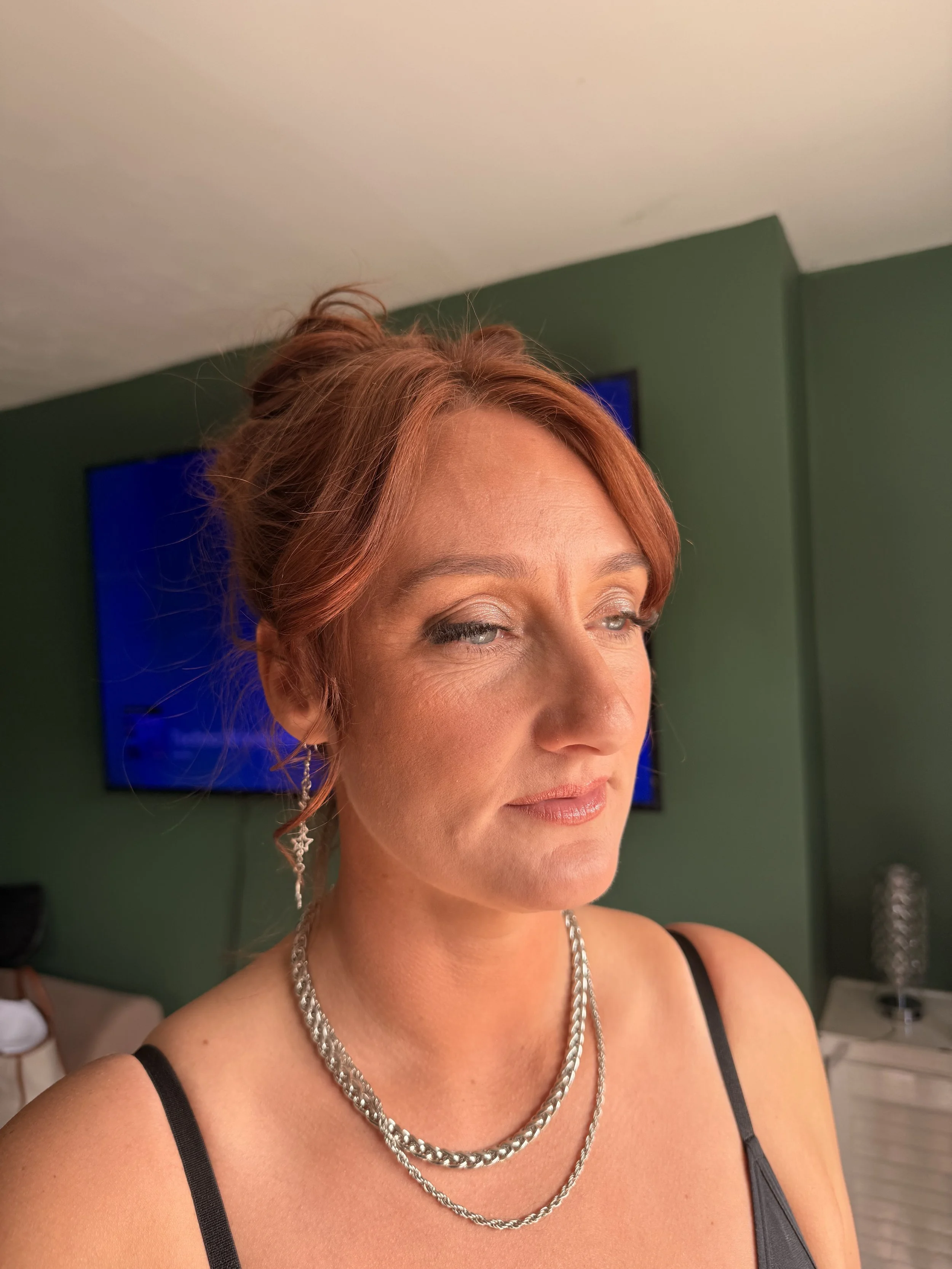 Close-up of a woman with red hair styled in an updo, wearing silver jewelry, and a black spaghetti strap dress, standing in a room with green walls and a television in the background.
