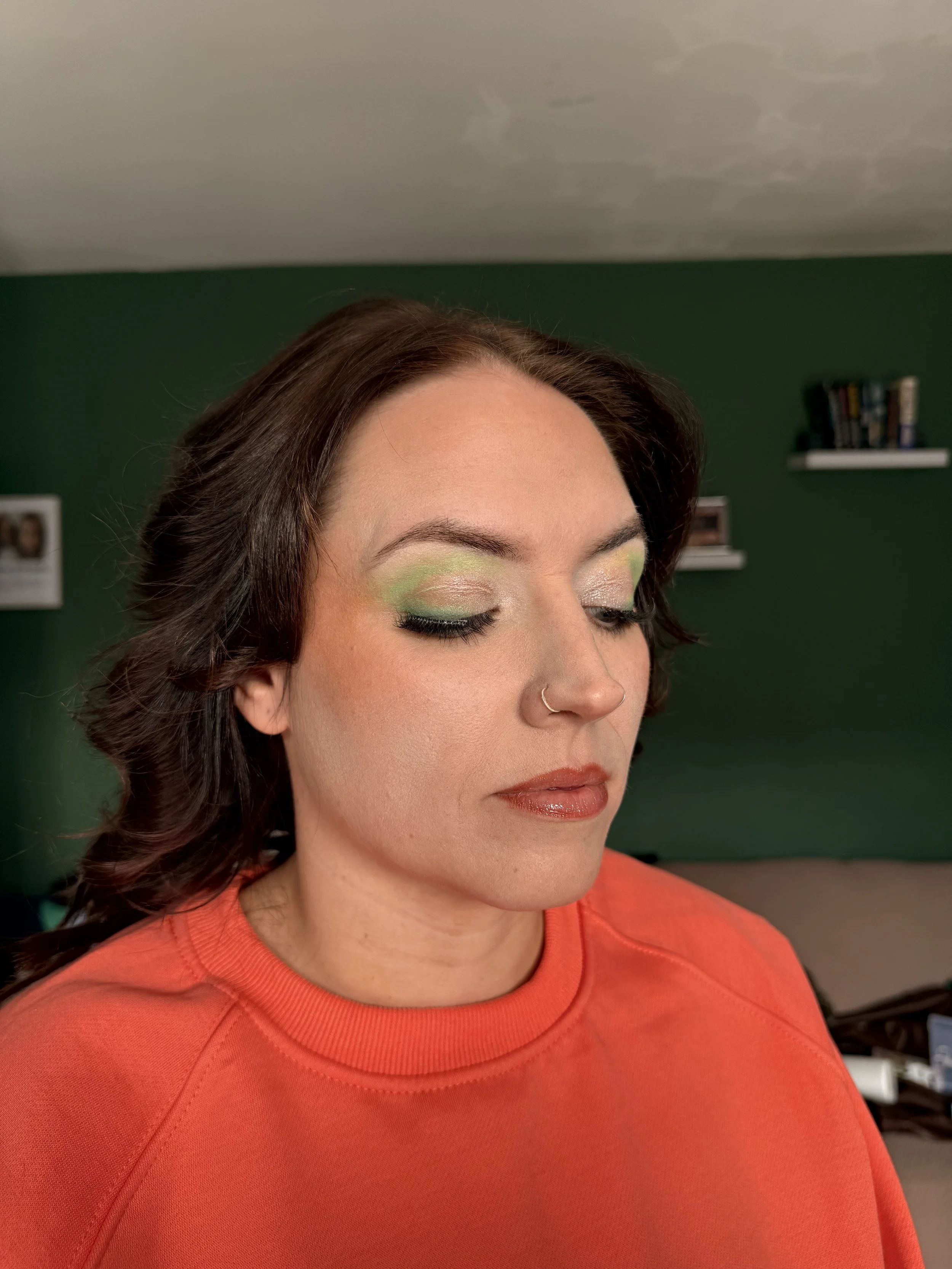 A woman with closed eyes and makeup, wearing a red top, in a room with green walls and shelves.