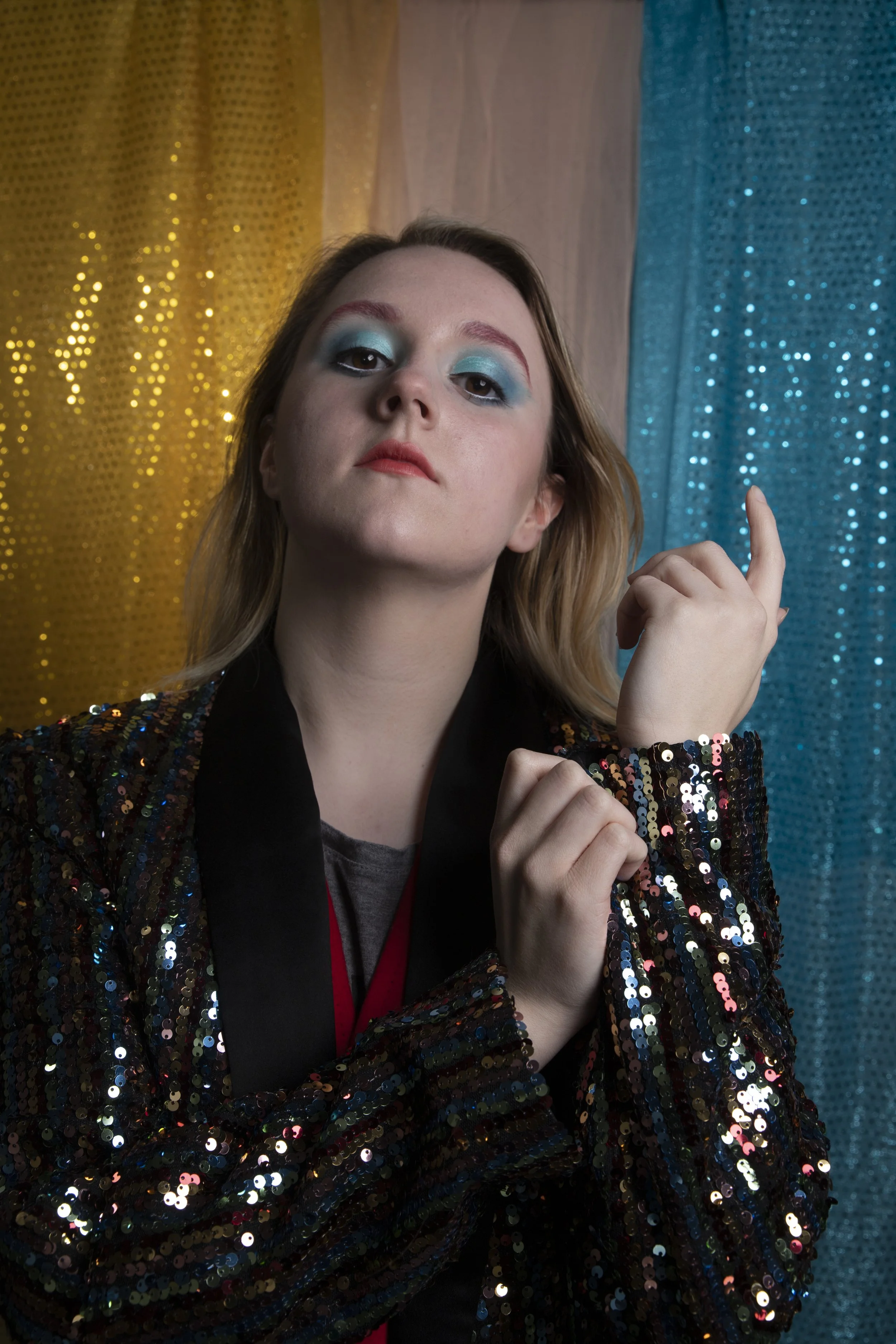 A woman with light skin and long, blond hair is wearing a black and colorful sequin jacket with a gray shirt underneath. She has makeup with blue eyeshadow, red eyebrows, and lipstick. She is pointing upward with her right hand and looking directly a