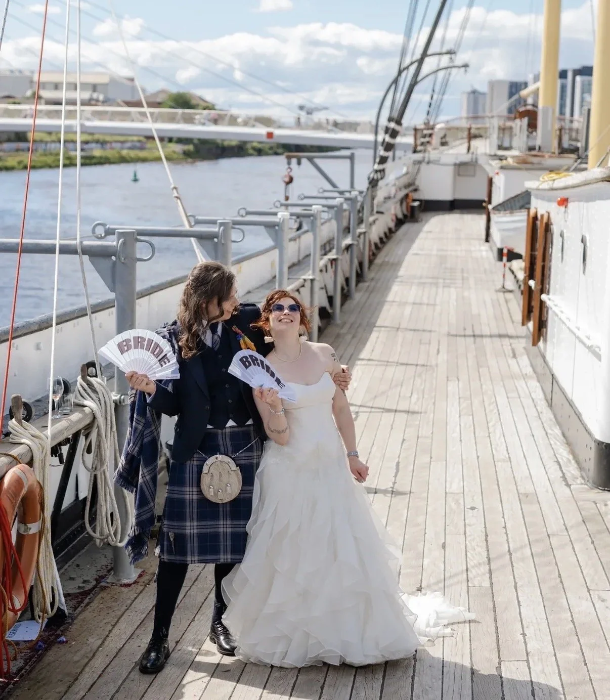 Two women dressed for a wedding standing on a ship's deck, holding fans that say 'BRIDE'. They are smiling and enjoying the moment with a body of water and city buildings in the background.