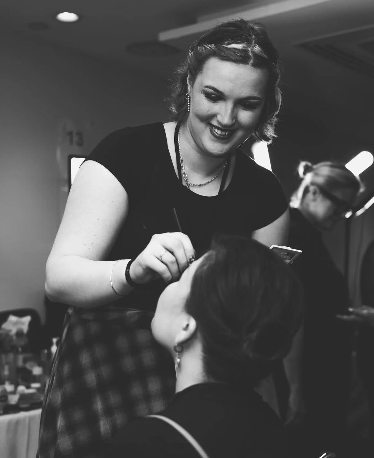 A woman with short, wavy hair and jewelry is smiling while applying makeup or eyeliner to another woman with long hair, who is sitting and looking up. The scene appears to be in a professional setting, possibly a salon or event.